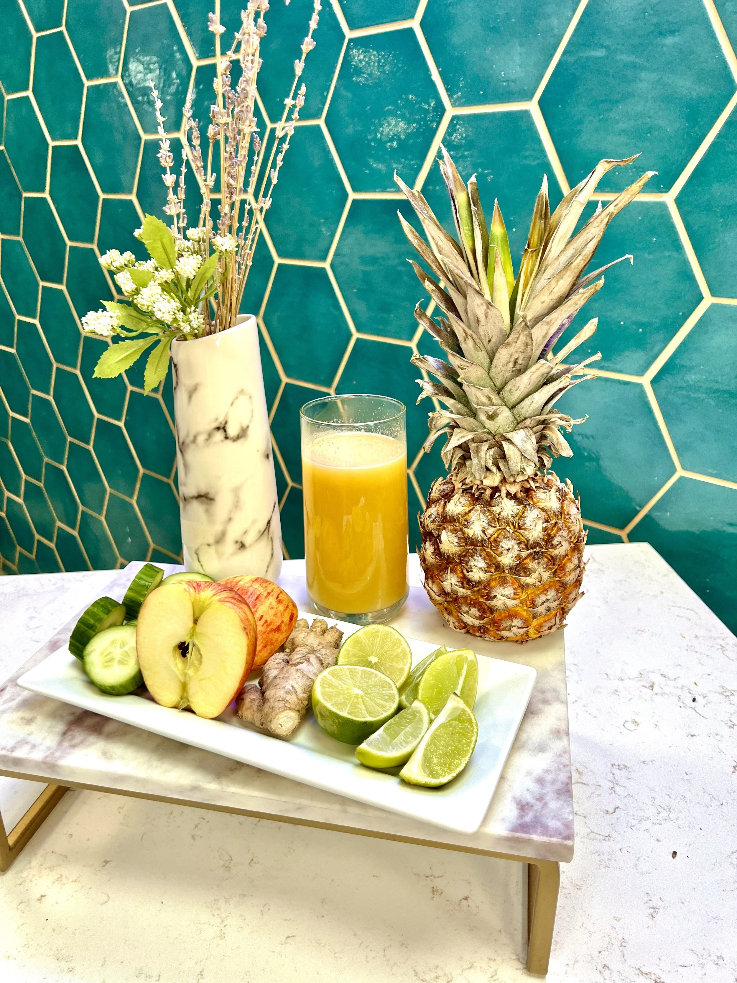A rectangular white tray with sliced cucumber, apple, ginger, and lime wedges next to a glass of orange juice and a whole pineapple. A vase with flowers is in the background, against a teal hexagonal tile wall.