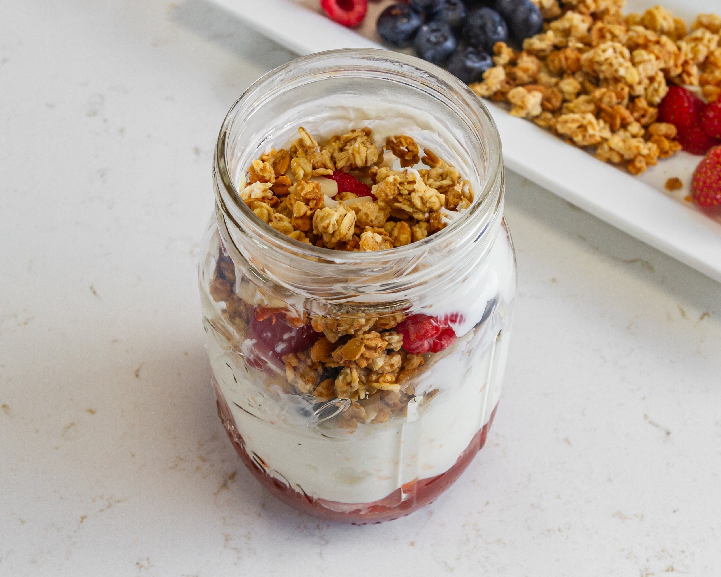 A glass jar filled with layers of yogurt, granola, and berries on a white surface.