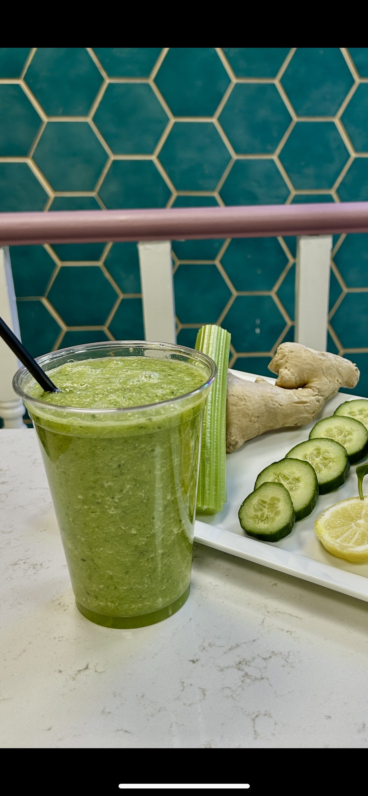 A green smoothie in a plastic cup with a black straw is on a table next to a plate of fresh ingredients, including celery sticks, ginger root, cucumber slices, and lemon slices. The background features a teal hexagonal tile pattern.