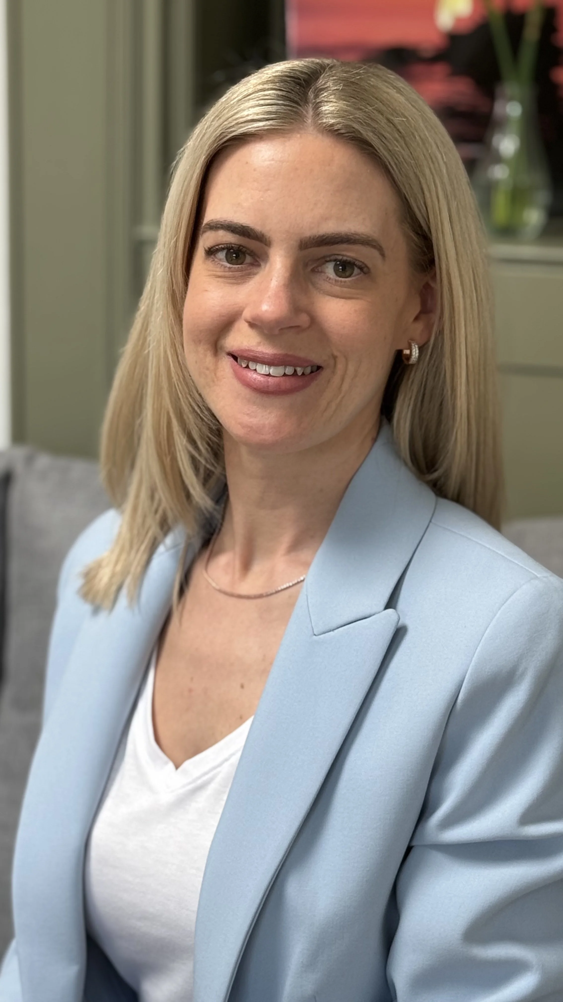 A woman with blonde hair, wearing a light blue blazer, white top, and jewelry, smiling in an indoor setting.
