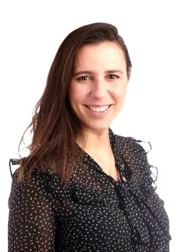 A woman with long brown hair smiling, wearing a black blouse with white polka dots, against a plain white background.