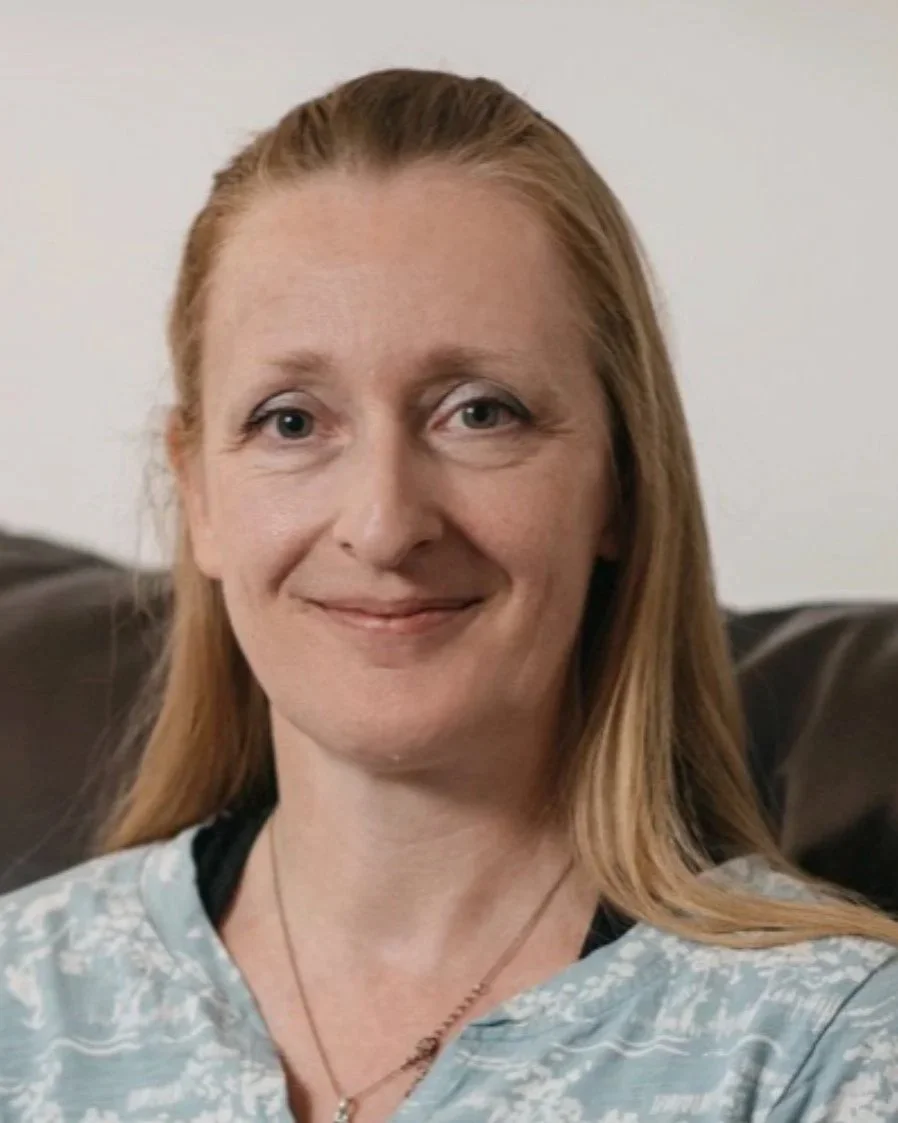 Close-up of a woman with red hair smiling, sitting on a dark brown sofa, wearing a light blue patterned top and a necklace.