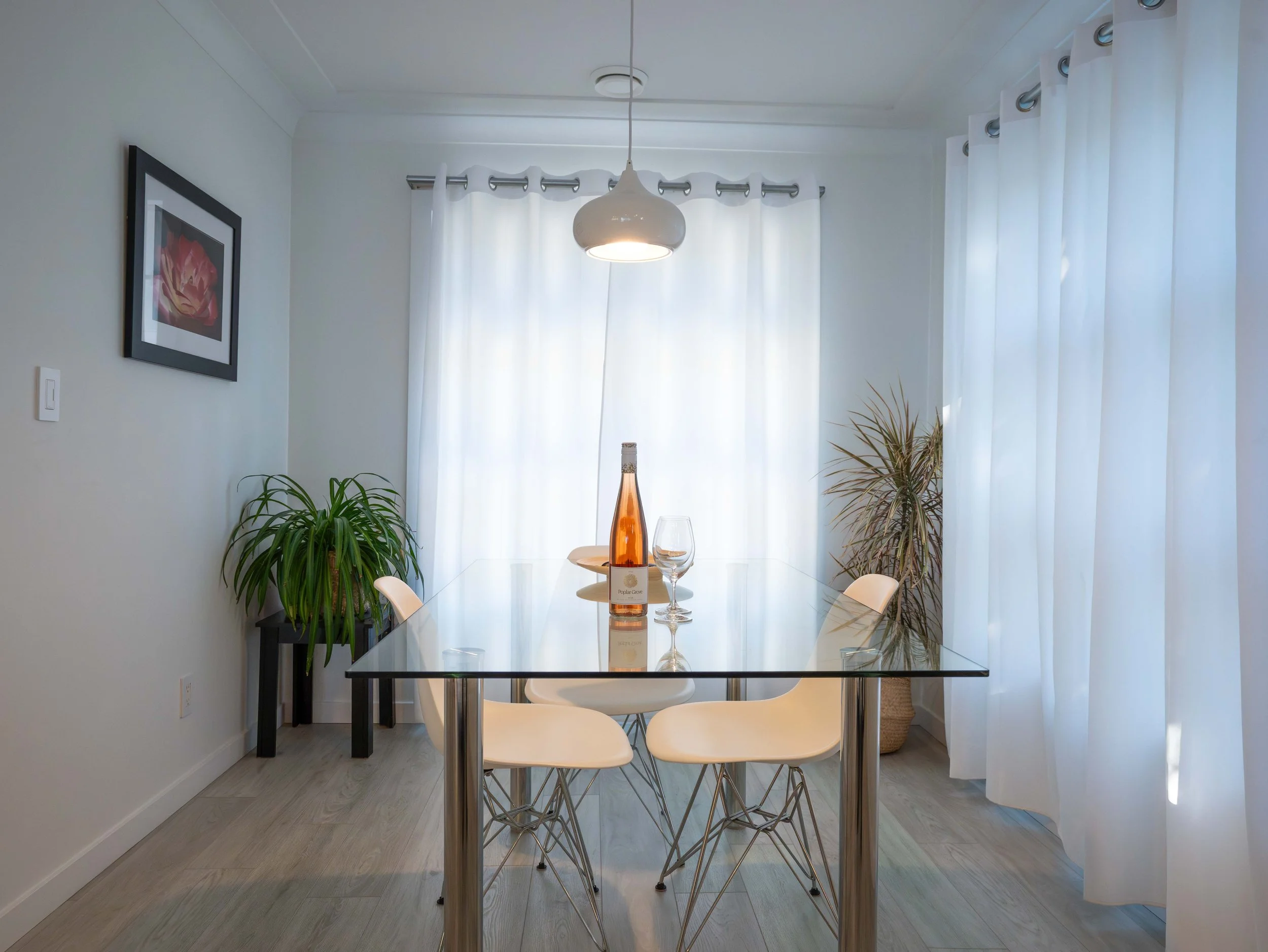 Modern dining room with glass table, white chairs, hanging light, and a bottle of Okanagan wine with glasses. Large white curtains and potted plants add decor. Beautiful interior dining room.