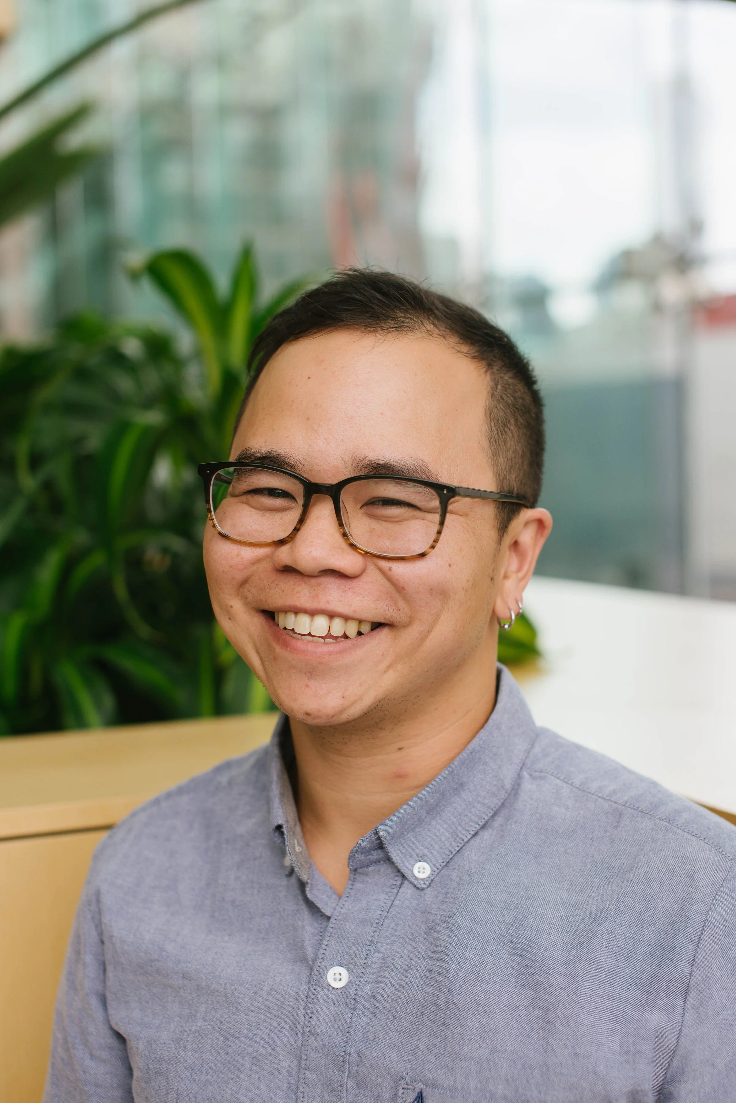 Smiling person wearing glasses and a light blue button-up shirt in a modern setting with plants in the background.