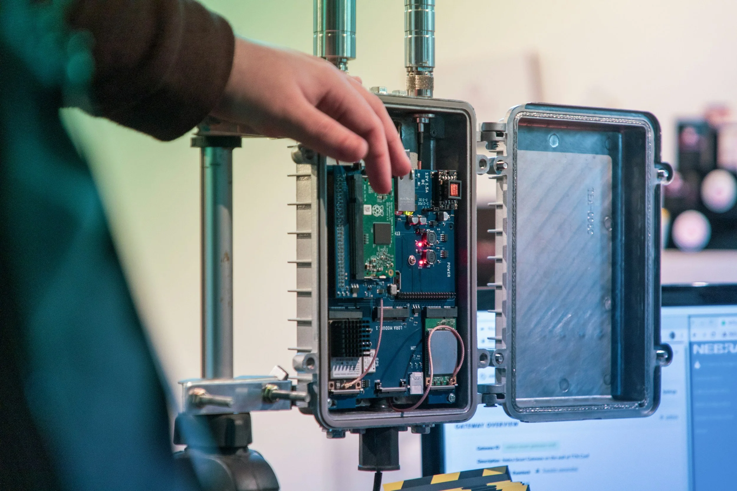 Close-up of a person's hand working on a circuit board and gateway inside a remote telemetry unit (RTU) enclosure, with a blurred background showing a computer screen.
