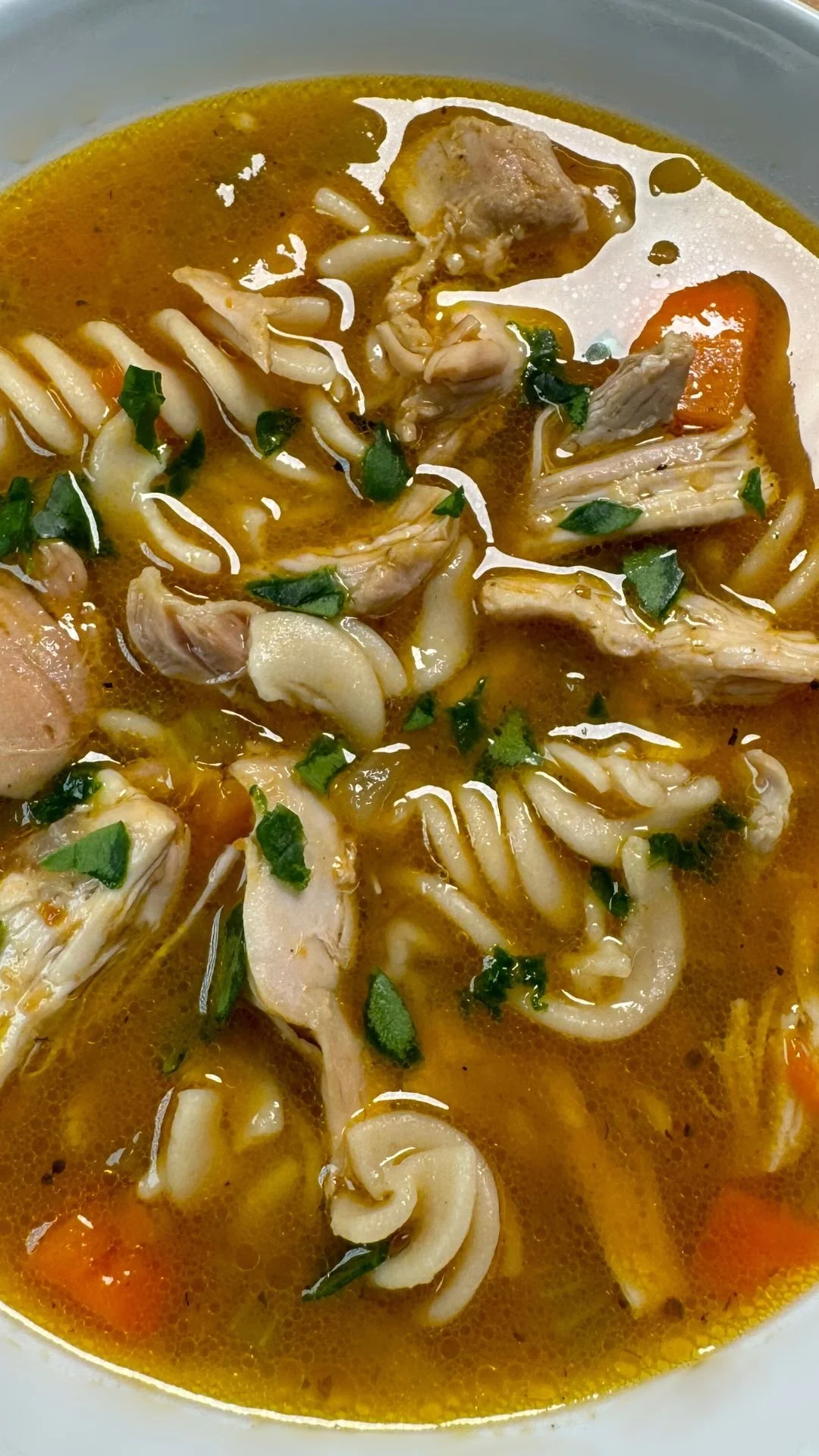Close-up of a bowl of chicken noodle soup with carrots, noodles, chicken pieces, herbs, and clear broth.