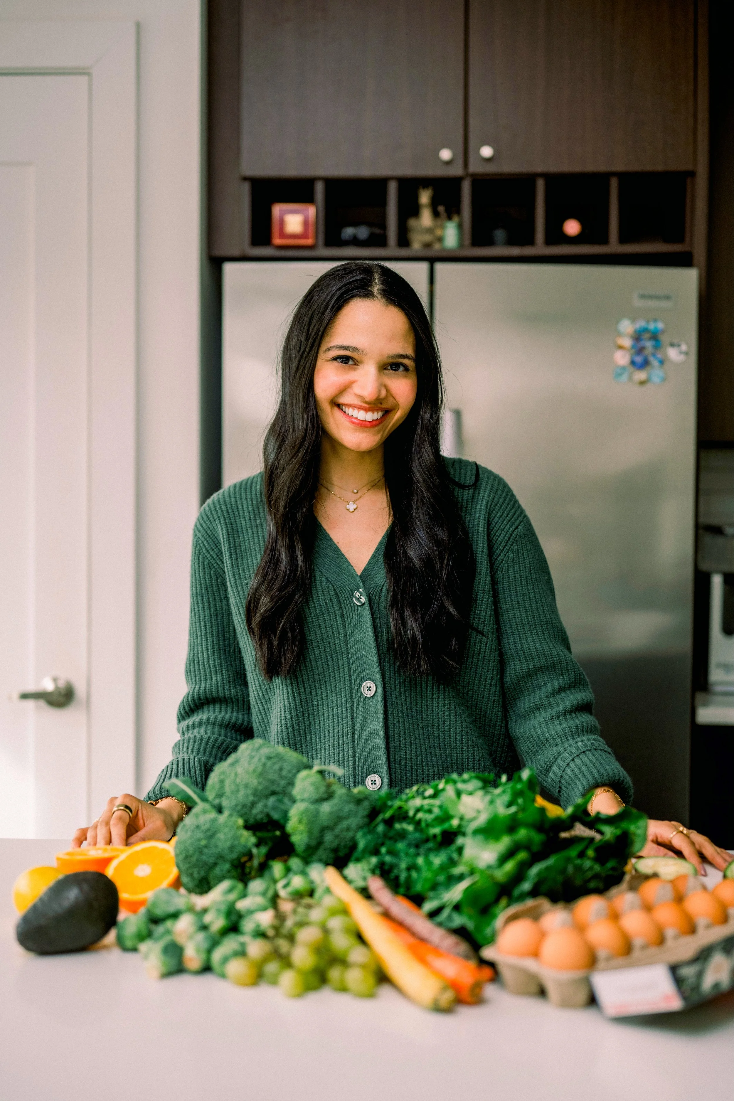 Woman smiling behind a variety of fresh vegetables and eggs on a kitchen counter.