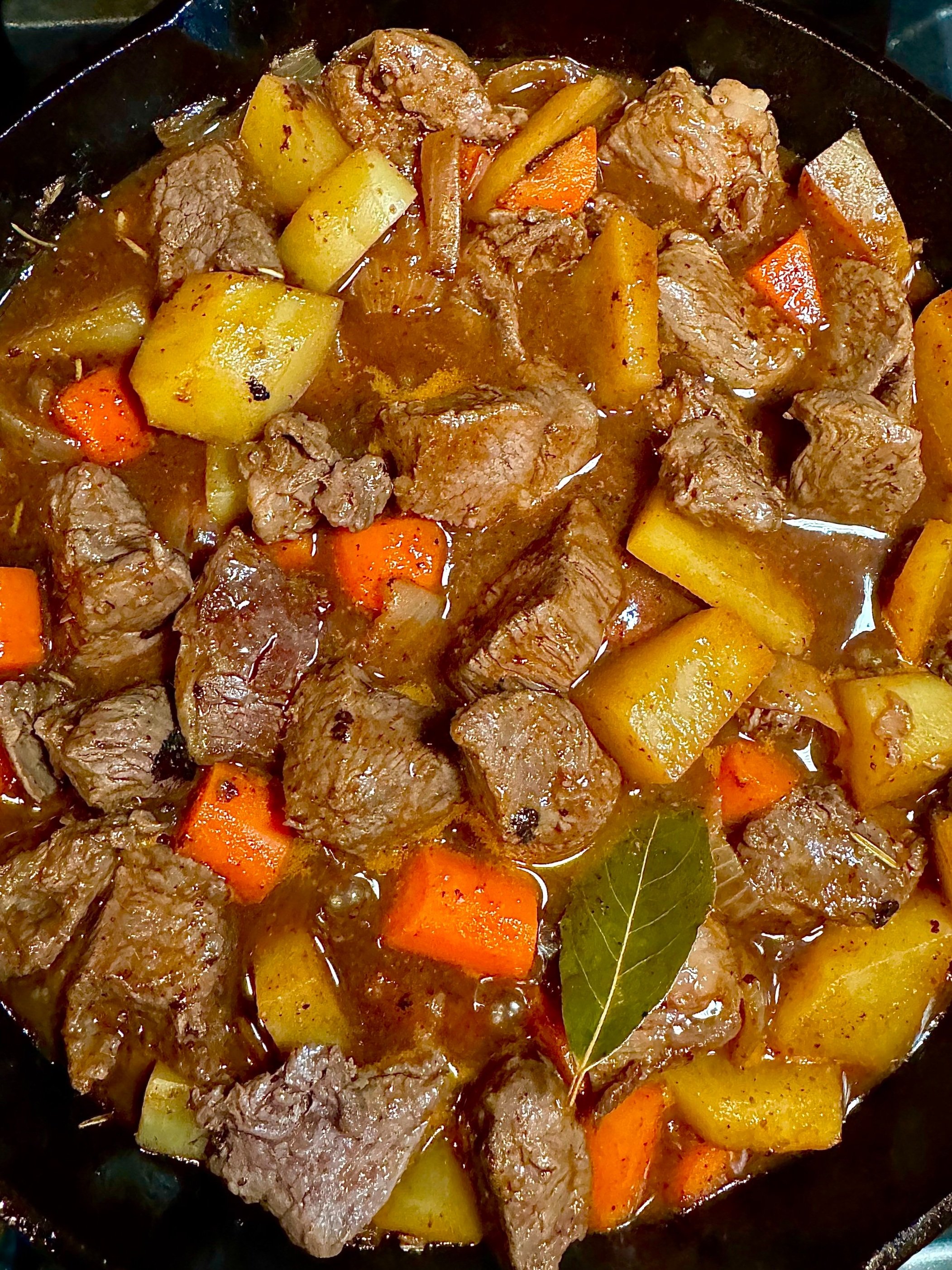 A close-up of a pot of beef stew with chunks of beef, potatoes, carrots, and onions, garnished with a bay leaf.