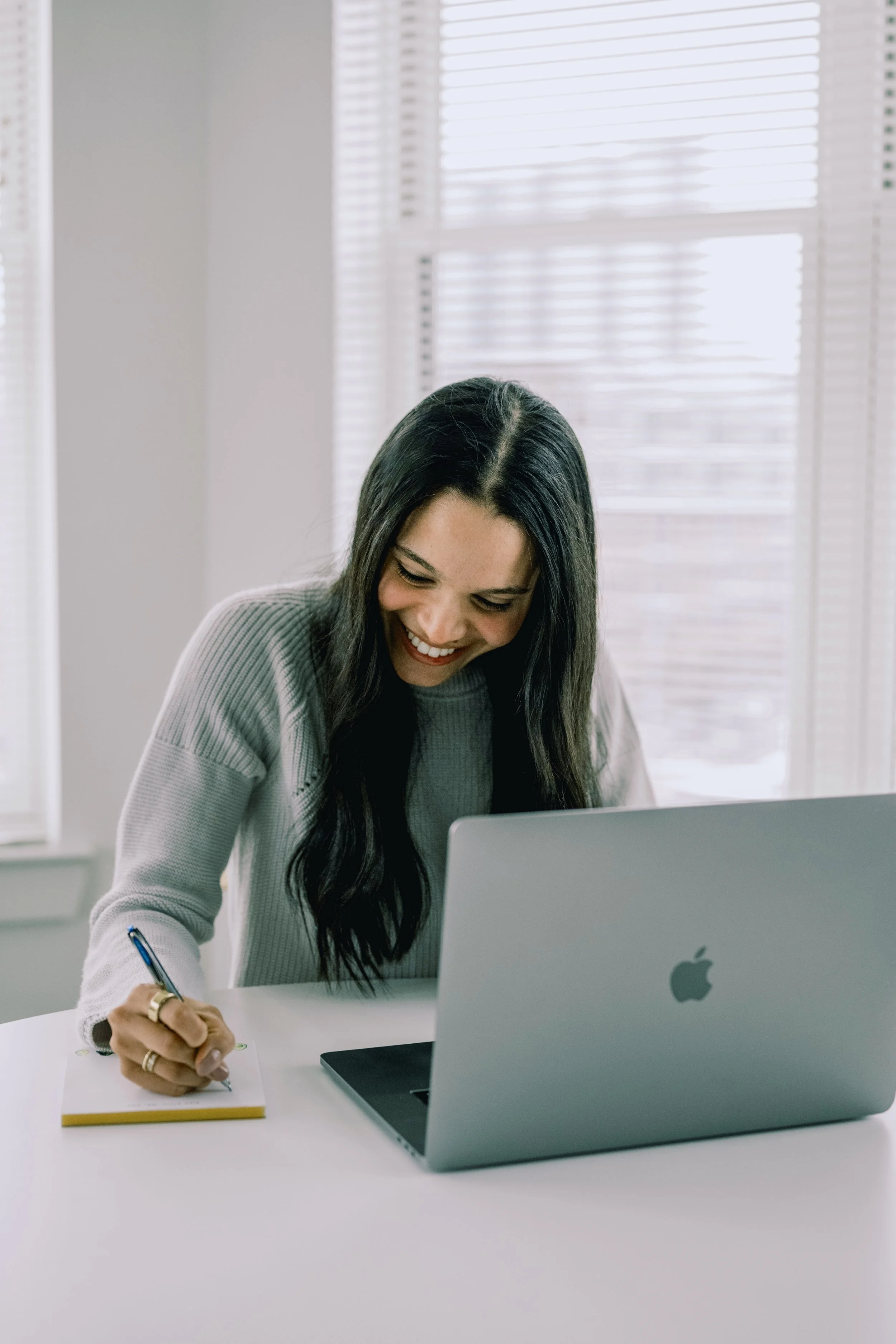 A woman with long dark hair smiling while working at a white desk with a silver Apple laptop, taking notes with a pen on a yellow notepad, in a bright room with large windows and blinds.