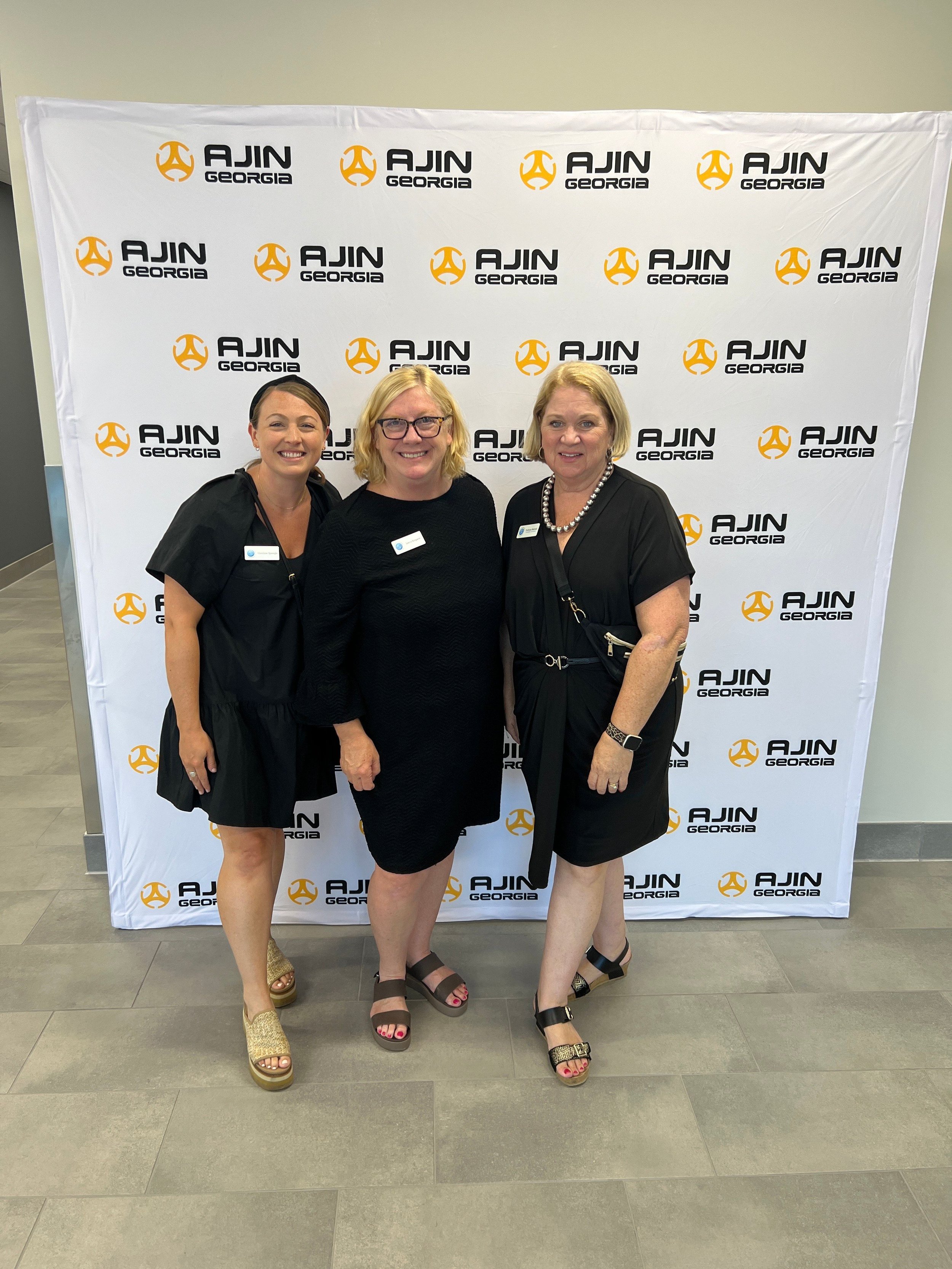 Three women in black dresses standing in front of a white backdrop with the logo and text 'AJIN Georgia' repeated, smiling at the camera.