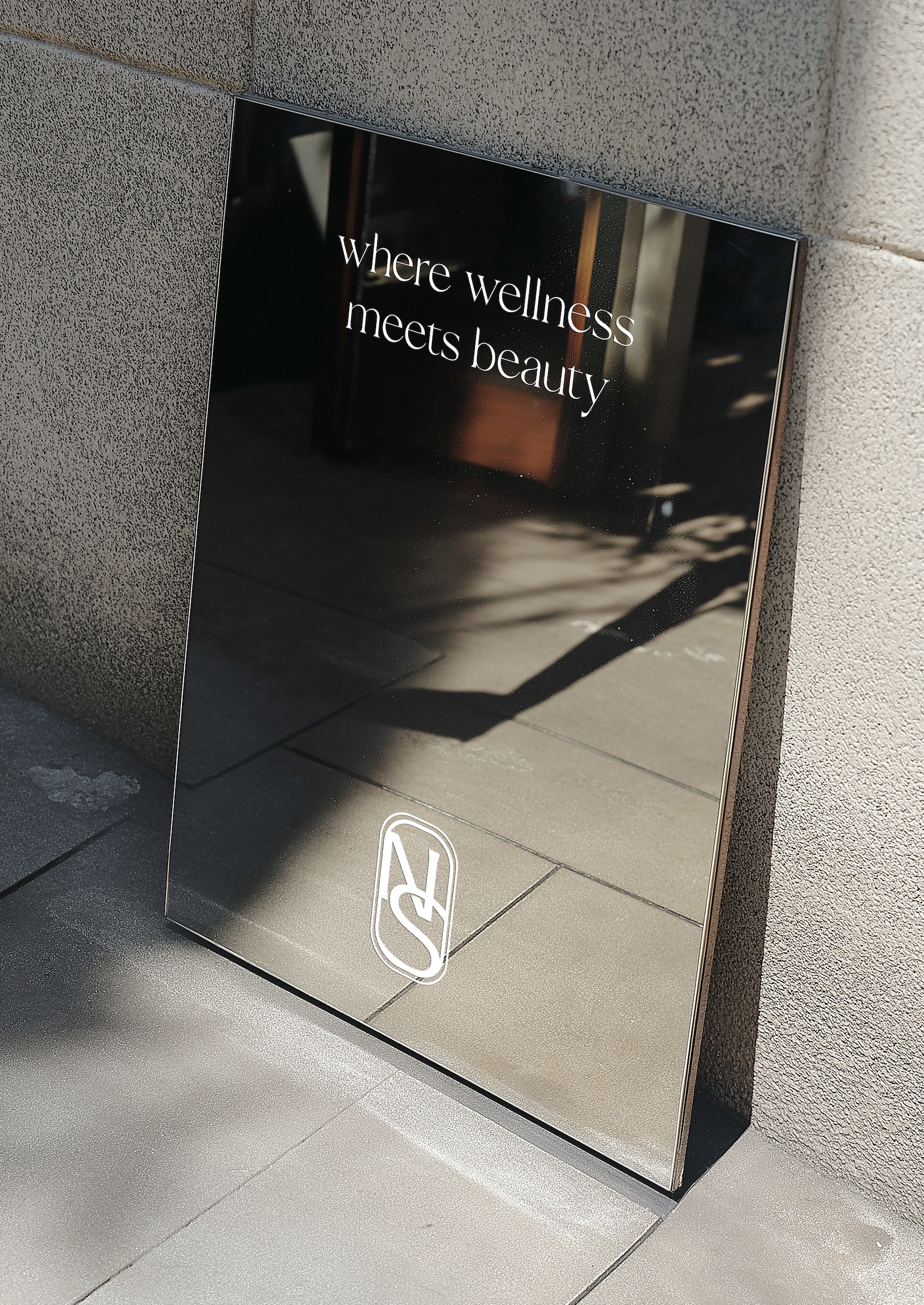 A reflective black sign with white text that reads "where wellness meets beauty" is mounted on a stone-textured wall. The sign has a logo in the bottom corner and shadows on the ground nearby.