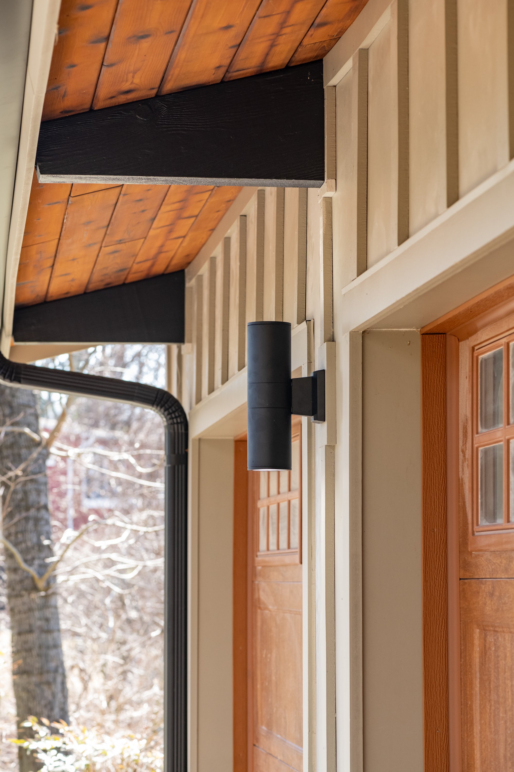 Exterior of a house with wooden siding, black modern wall light, wooden ceiling, and rain gutter system.