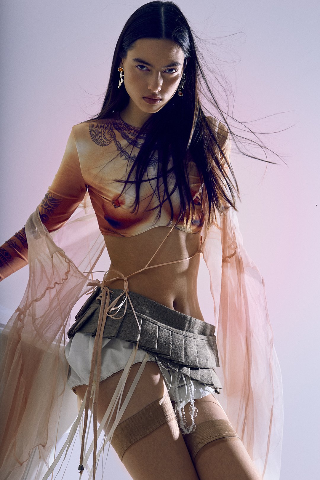 A woman with long dark hair wearing a sheer pink cover-up, a tan crop top, beige shorts, and thigh-high stockings, standing against a plain background.