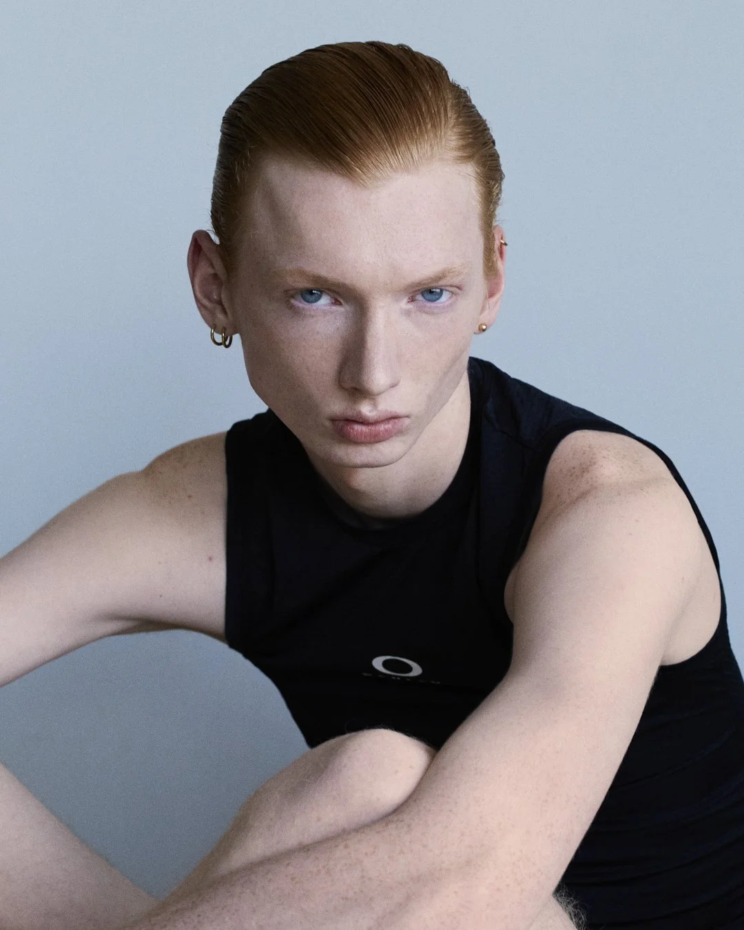 A youthful person with red hair, fair skin, and blue eyes wearing a black sleeveless top, looking directly at the camera with a serious expression.