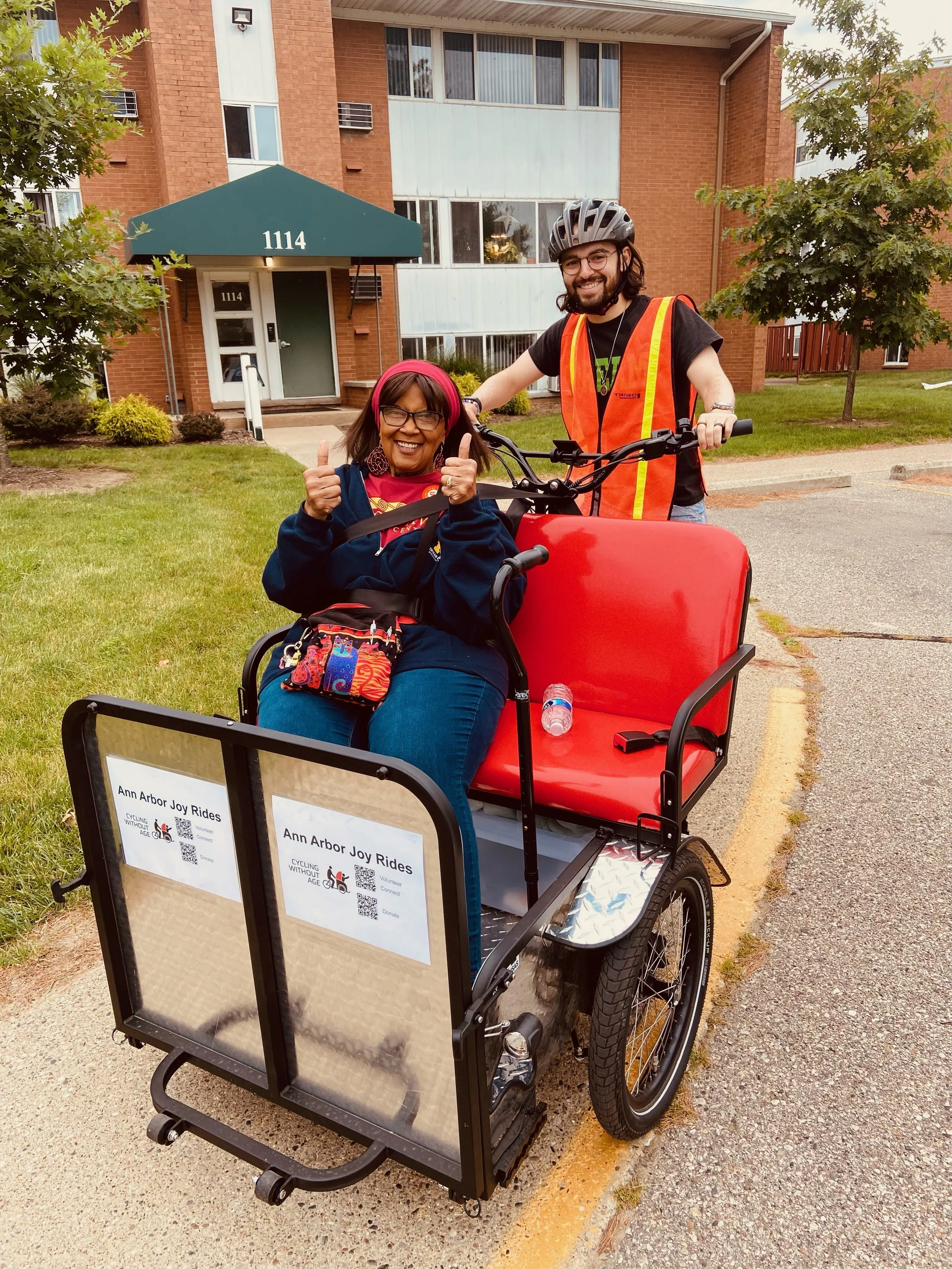 Volunteer Dylan pedals trishaw with co-pilot Dee smiling with thumbs up in front seat of bike