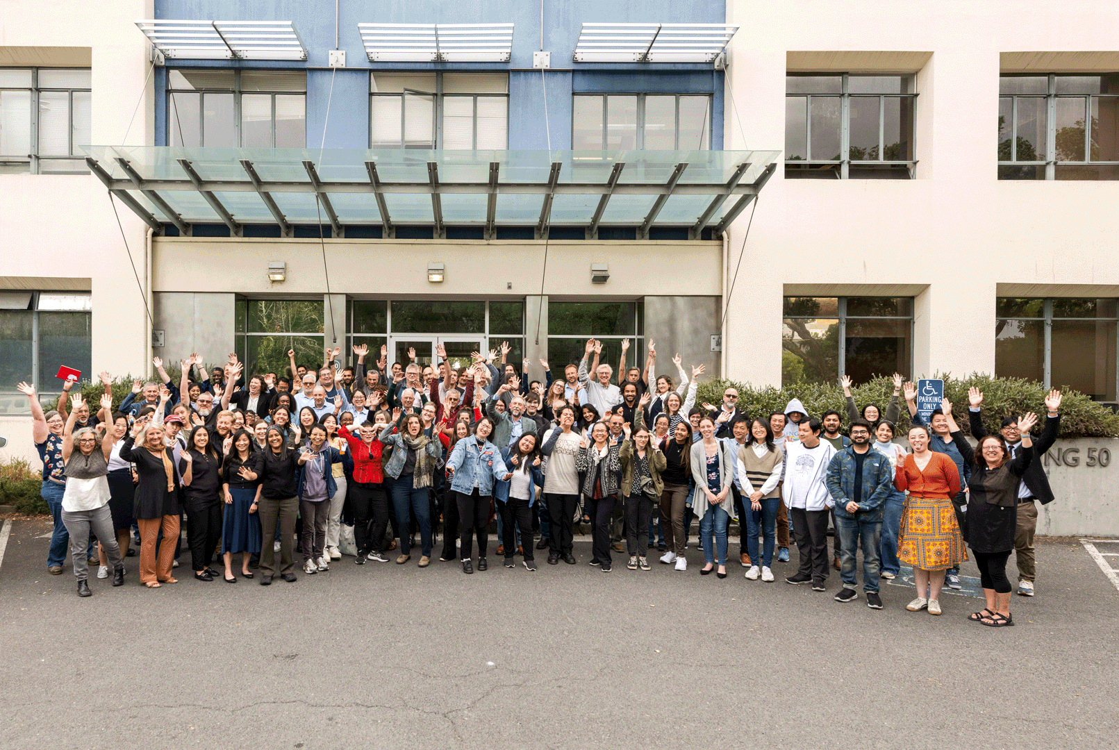 A large group of people posing in front of a building, with many raising their hands cheerfully.