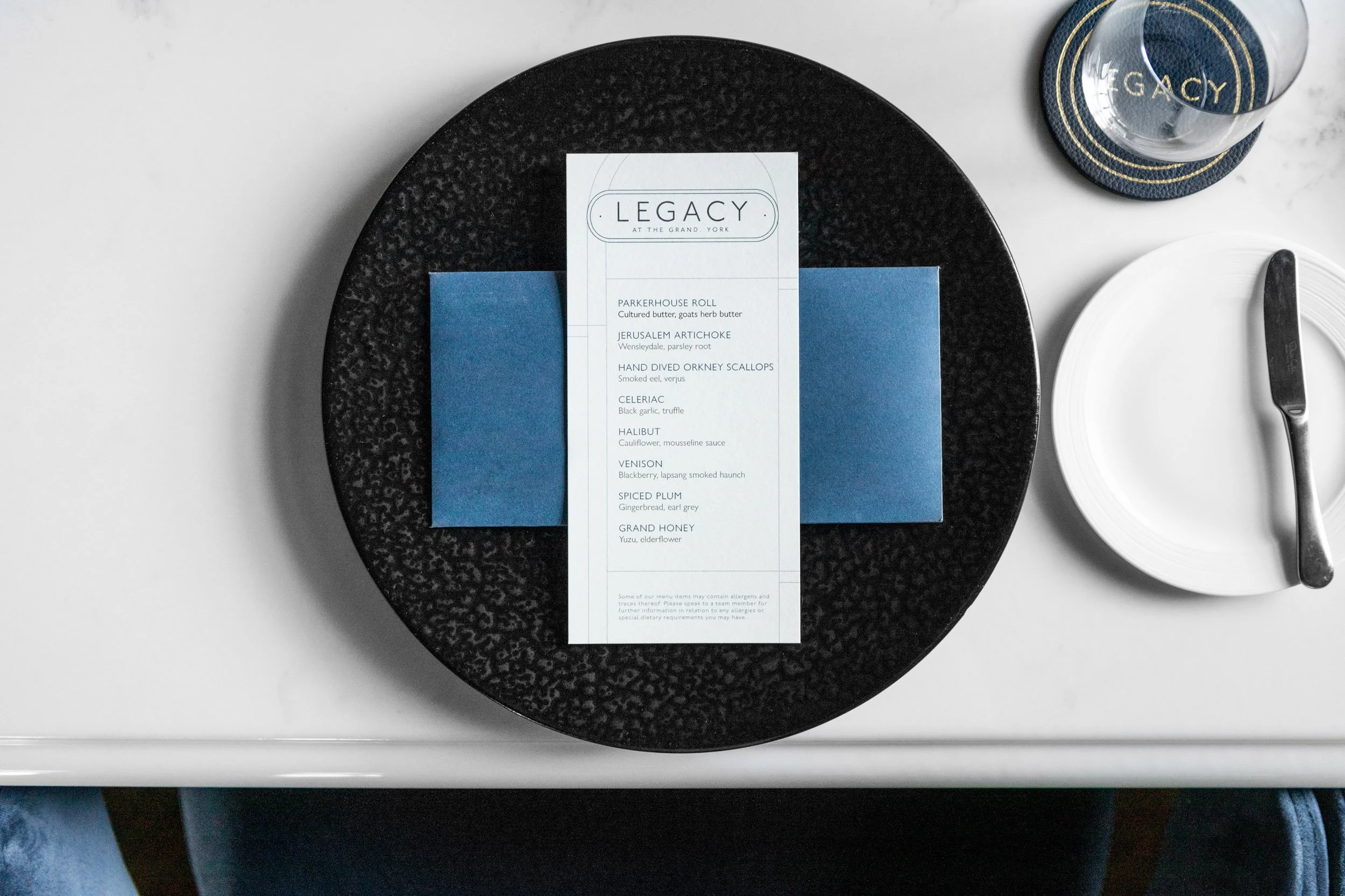 A black textured plate with a white and blue menu from Legacy at The Grand York, listing various gourmet food items, beside a white plate with a silver butter knife, a water glass on a blue coaster, and a white marble table surface.