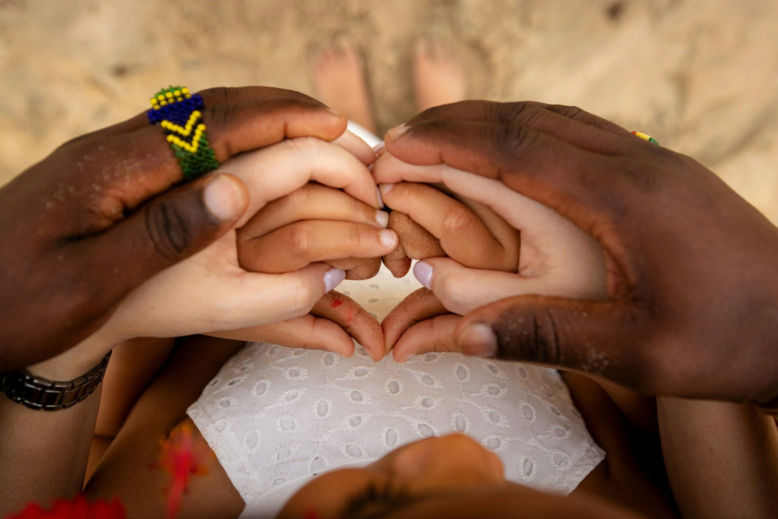 Multiple hands forming a heart shape over a person sitting on a sandy beach, wearing a white eyelet dress and colorful beaded bracelets. A multiracial family experiencing love.