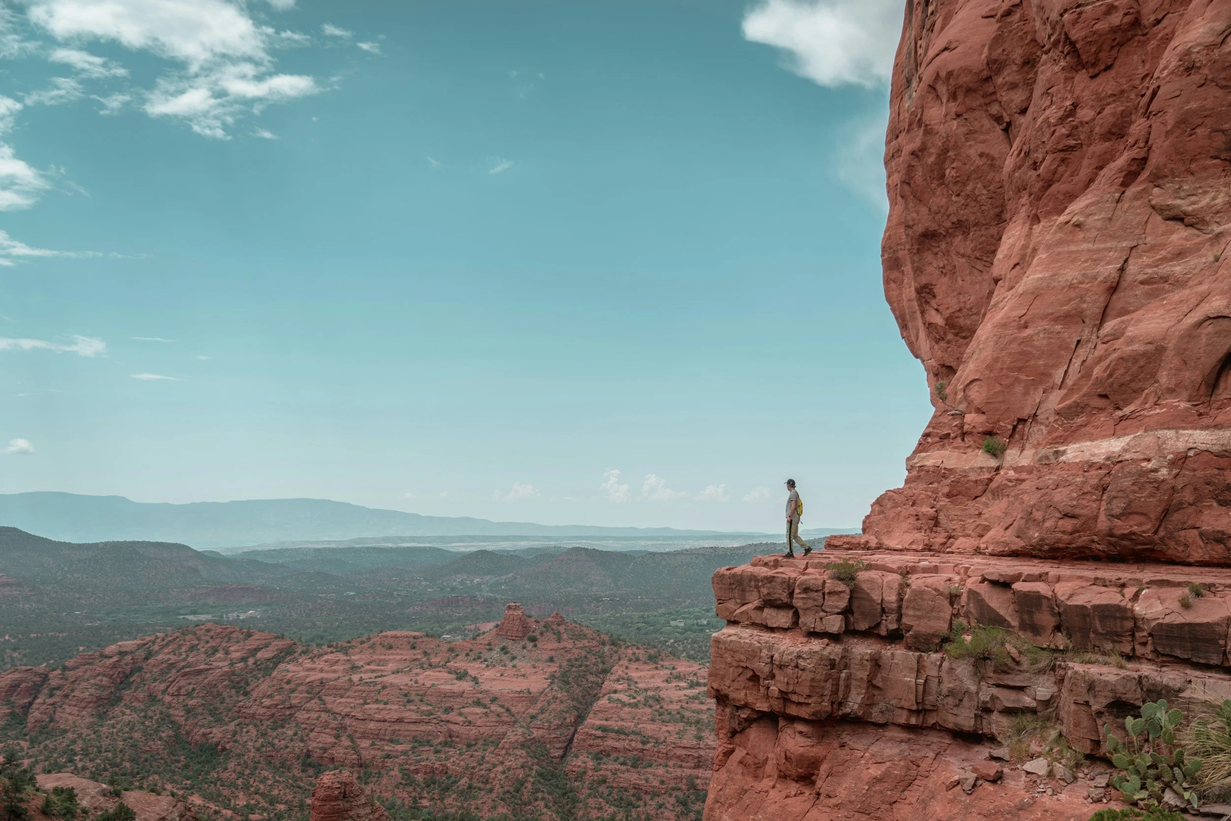Person standing on a rocky ledge near a red sandstone cliff with a desert landscape in the background under a cloudy sky.
