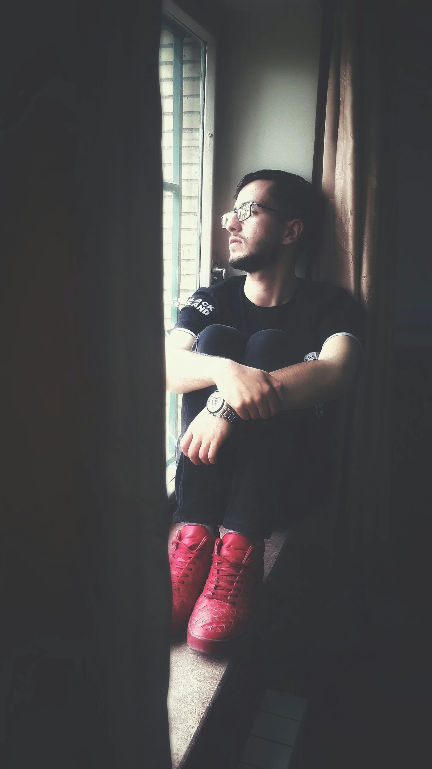 A young man sitting by a window, wearing black clothing, red shoes, glasses, and a watch, looking outside with a contemplative gong to therapy in Los Feliz.