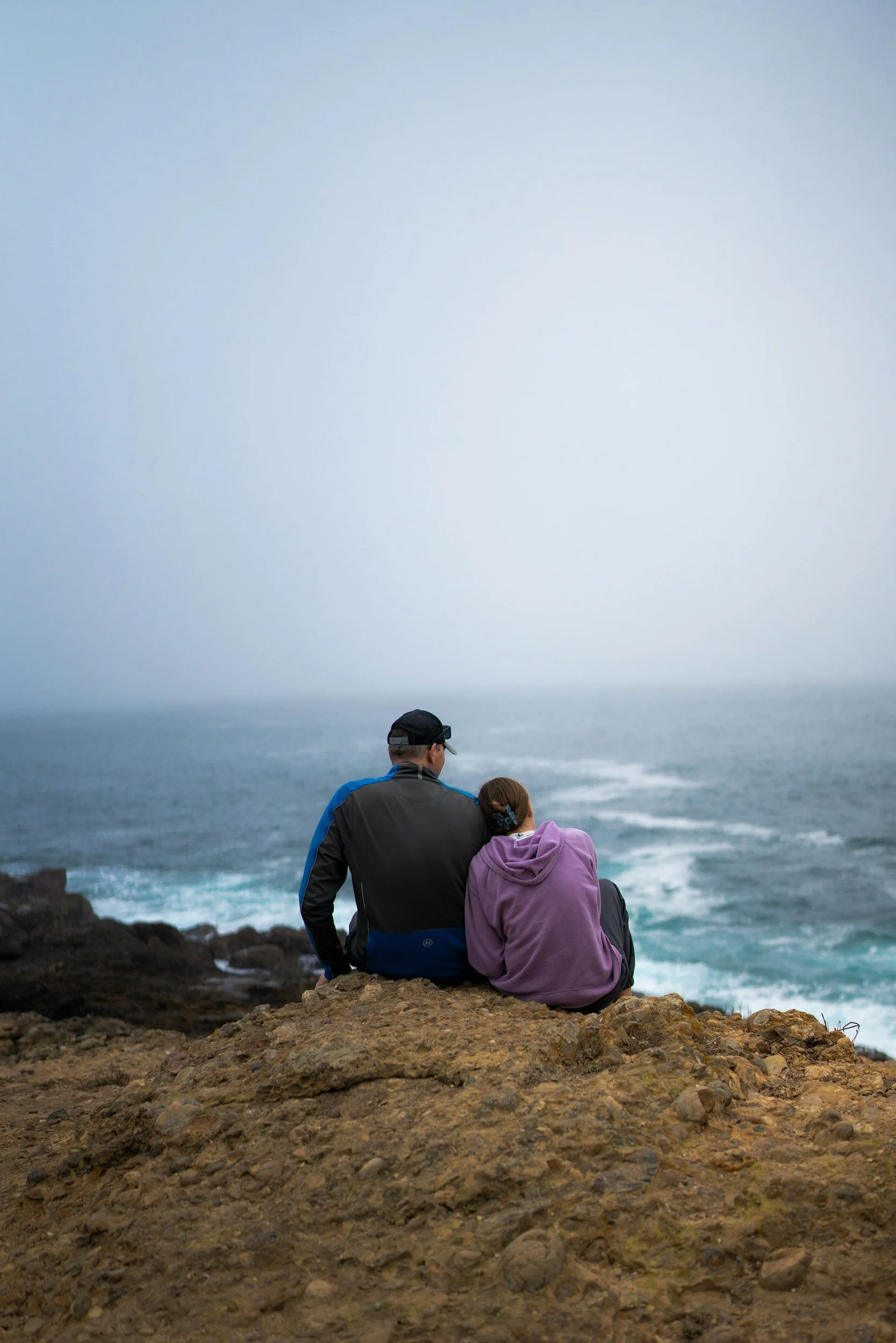 A couple sitting on a cliff overlooking the ocean, woman leaning her head on man's shoulder after a therapy session in Santa Barbara.