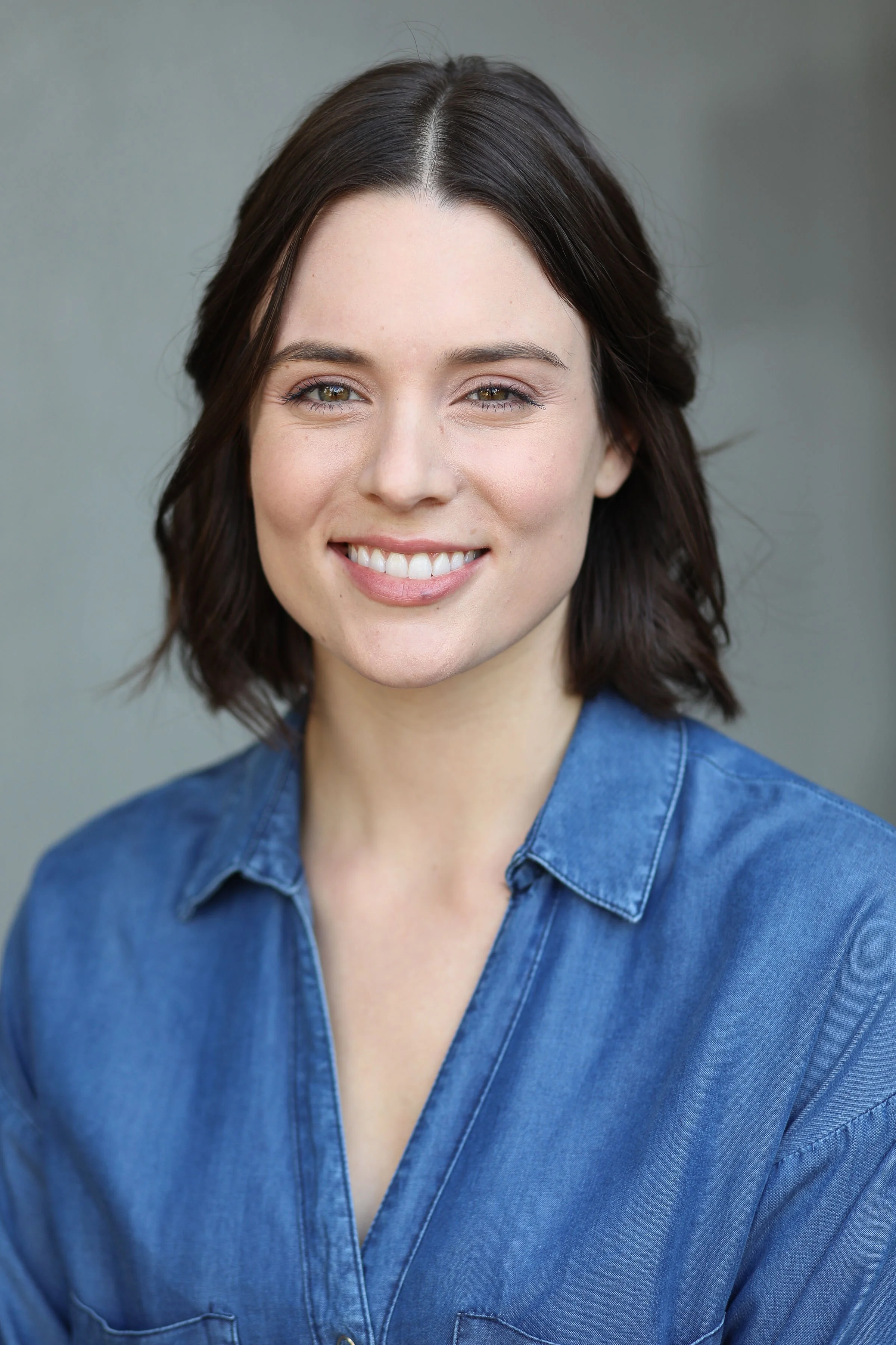 A young female therapist with short brown hair, wearing a denim shirt, smiling at the camera with a gray background.