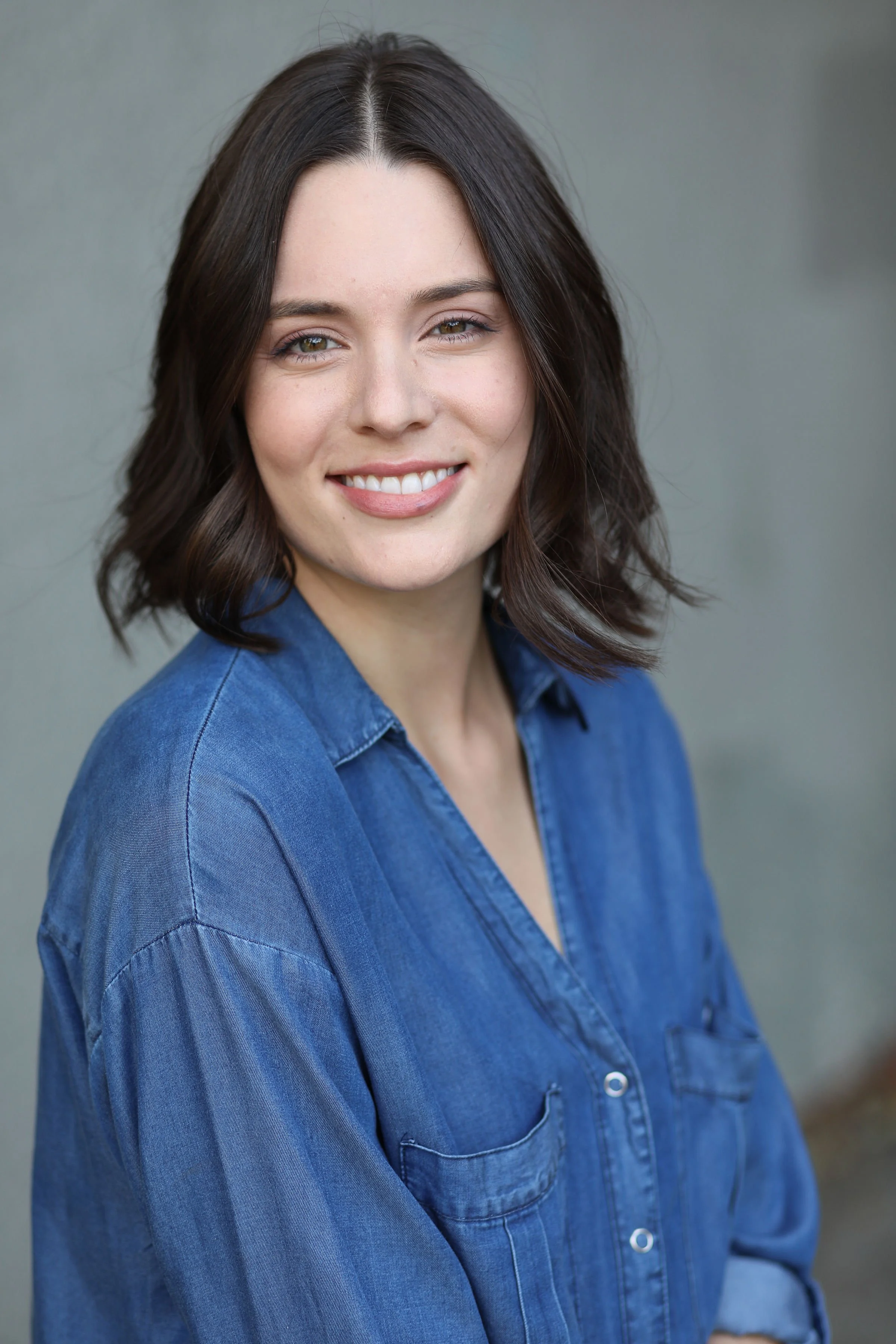 A female marriage and family therapist with shoulder-length dark brown hair, smiling, wearing a blue denim shirt, standing outdoors against a gray background in Los Angeles, CA.