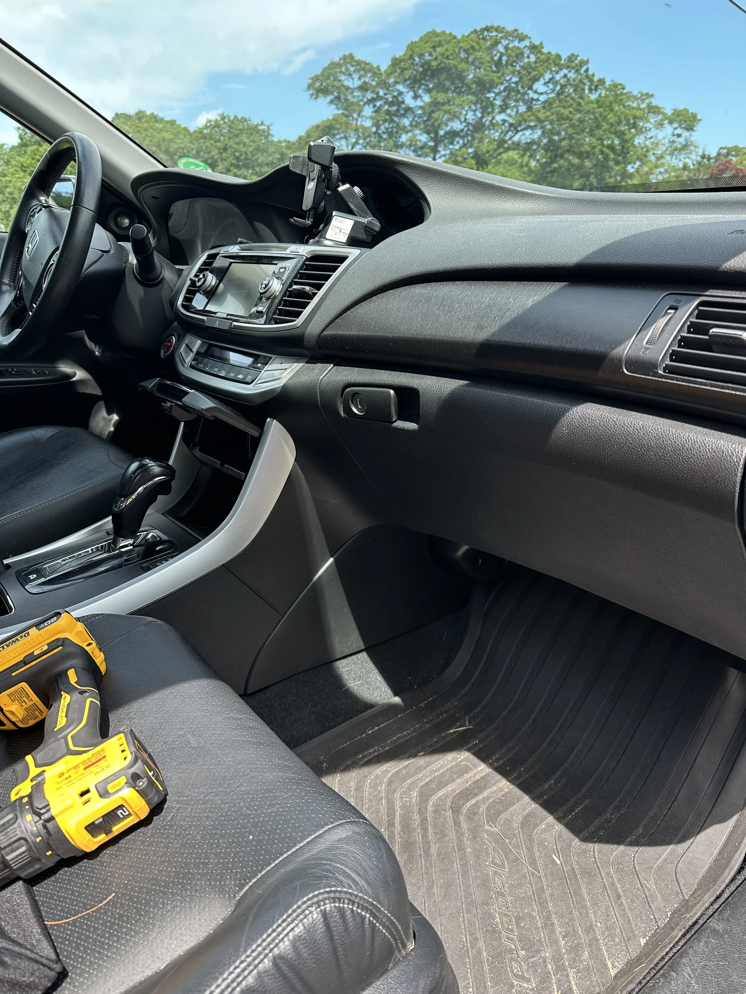Inside the front passenger side of a vehicle, showing a dashboard with an infotainment screen, a gear shift, and tools on the seat, with trees and a blue sky visible through the windshield.