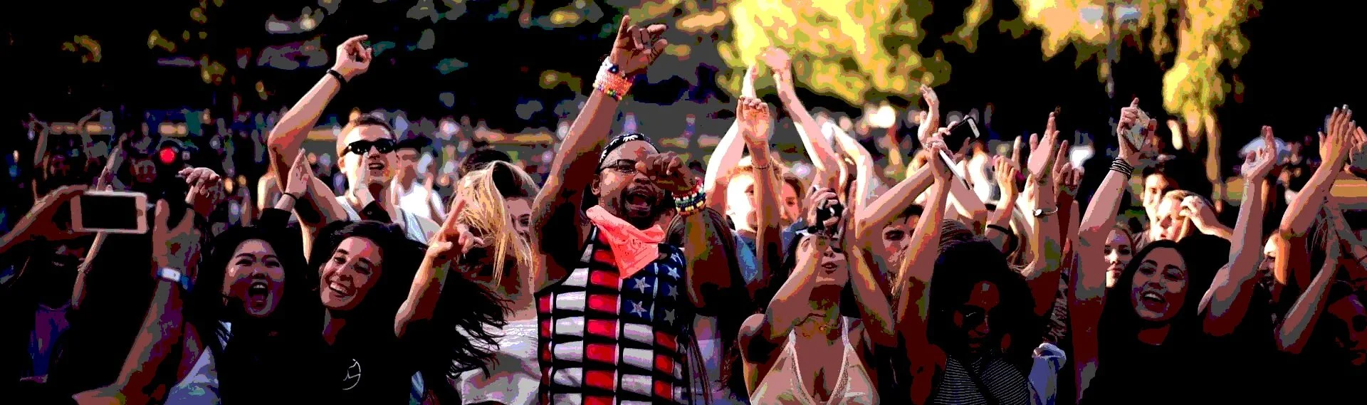 A crowd of people at an outdoor concert, dancing and singing, with some holding up phones, under a backdrop of trees and sunlight.