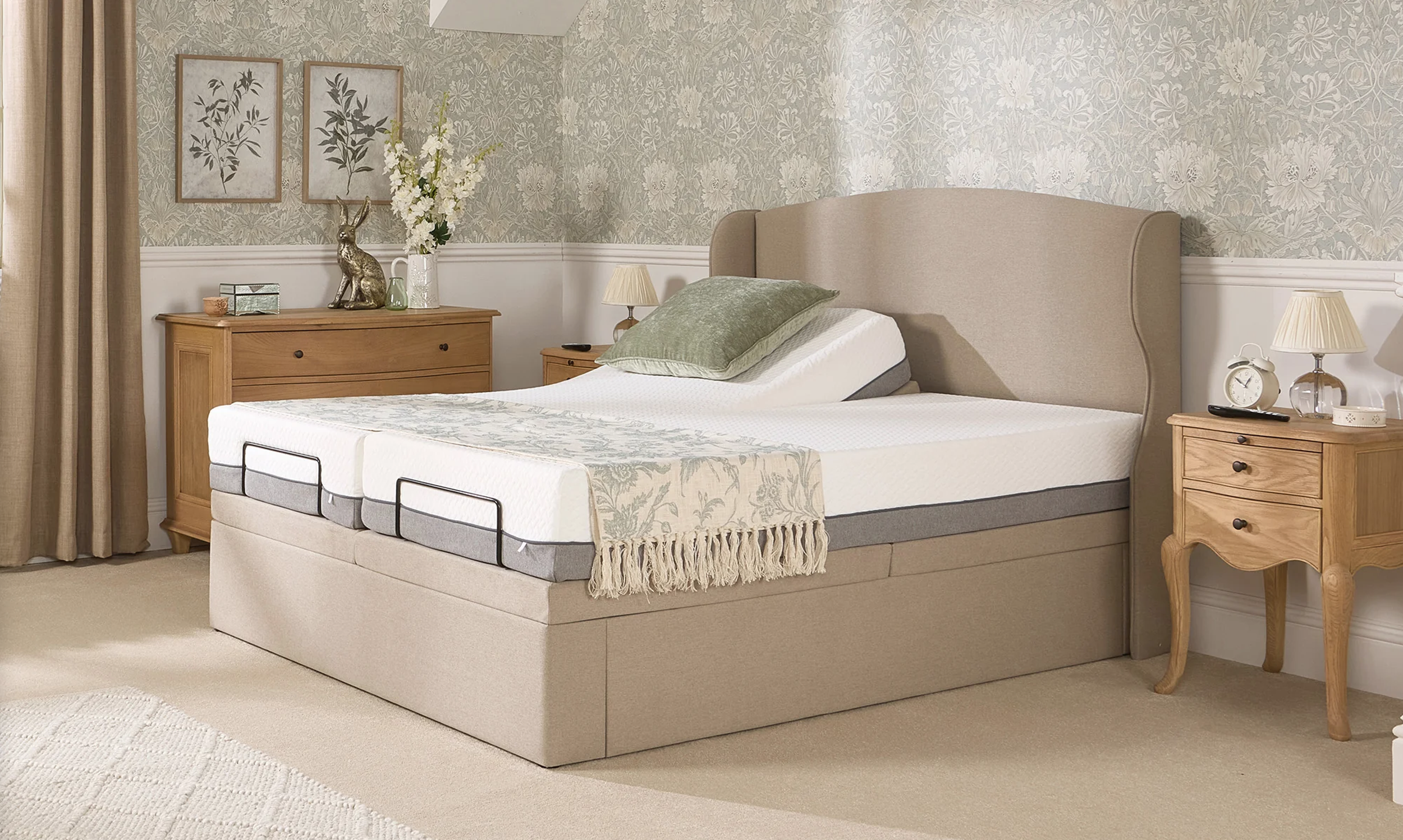 A bedroom with a beige upholstered bed, green pillow, floral throw, and wooden nightstands with lamps, a clock, and decorative items, with floral wallpaper and curtains.