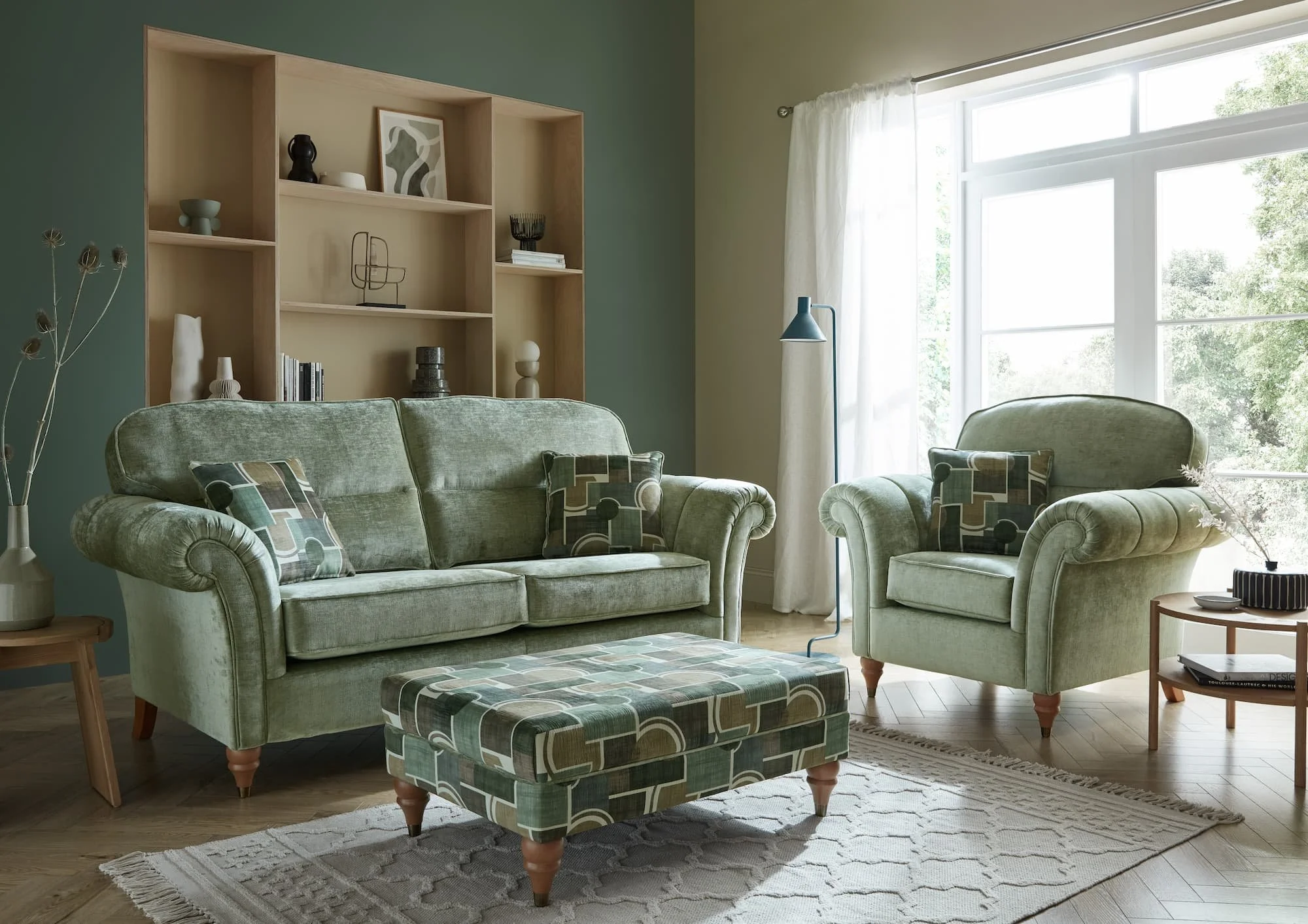 Living room with green upholstered sofa and armchair, patterned ottoman, wooden side tables, bookshelf with decorative items, large window with white curtains, and a green wall.