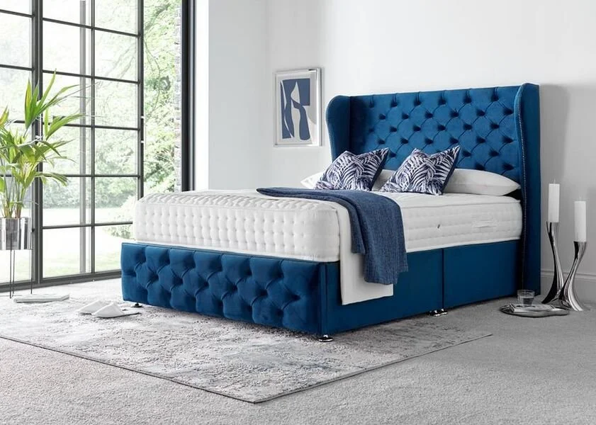 A modern bedroom with a blue velvet tufted bed, white mattress, and decorative pillows, next to a large window and abstract artwork, with a gray rug and candles on the side.