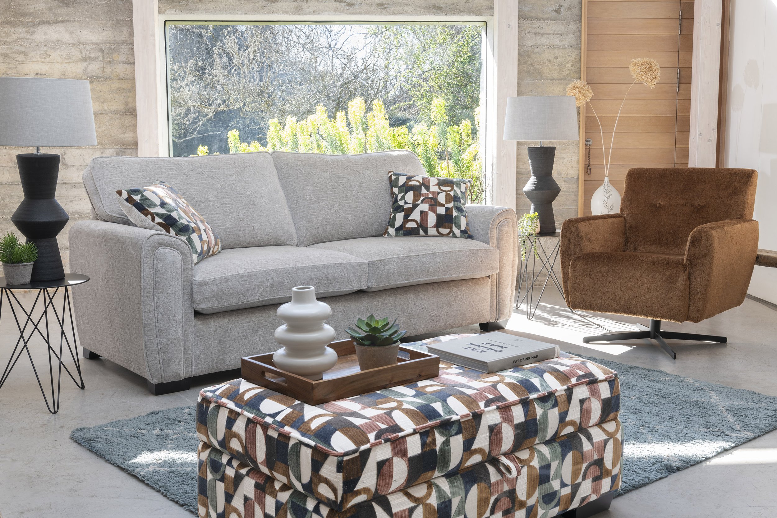 Living room with a light gray sofa, a patterned ottoman, a brown armchair, two side tables with lamps, and decor, overlooking a large window with green trees outside.