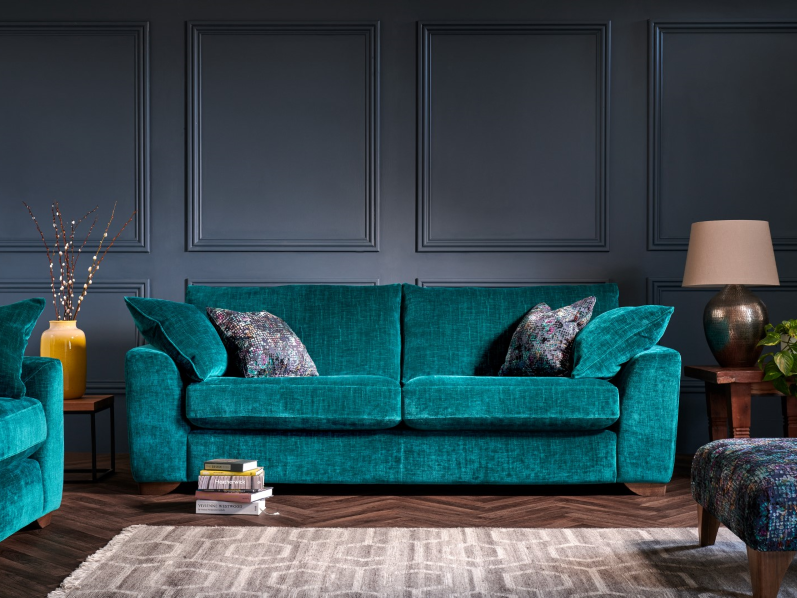 A teal sofa with two decorative pillows in a living room with dark blue paneled walls, a wooden side table with a lamp, a small yellow vase with decorative branches, a stack of books, an armchair, and a patterned ottoman.