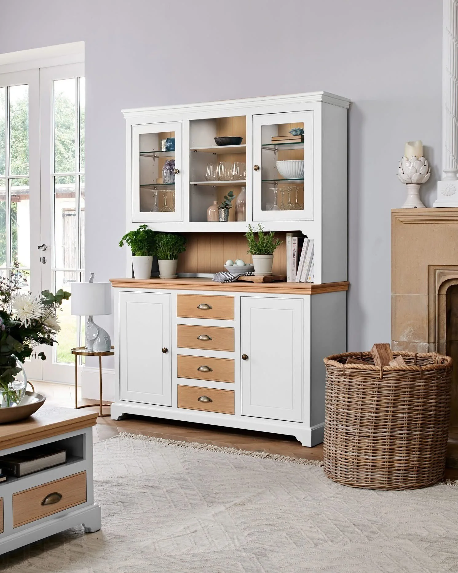 A white and wood-colored china cabinet with glass doors in a living room, next to a fireplace and a wicker basket, with a glass-door patio and window in the background.
