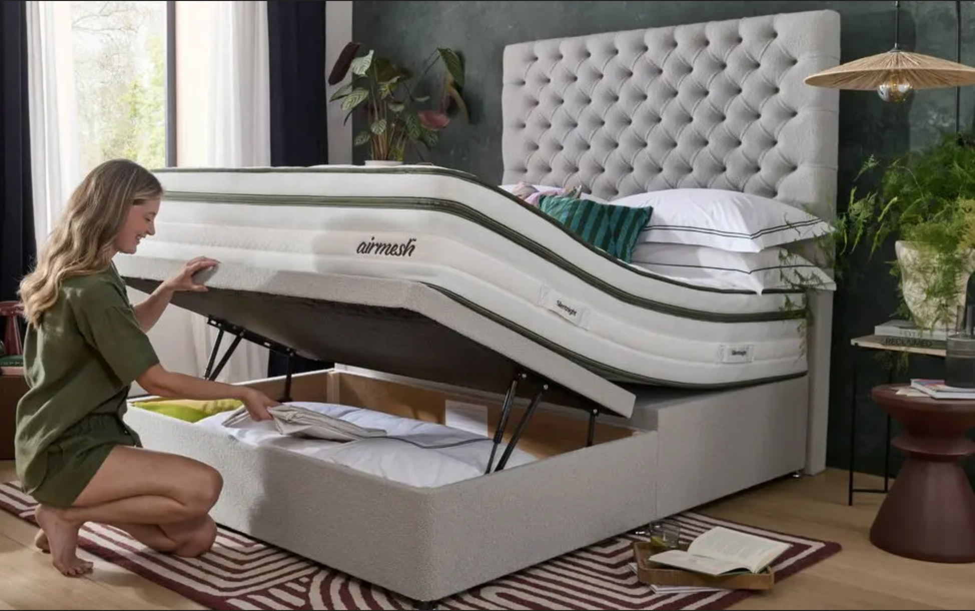 A woman opens a storage compartment under a modern bed with an advanced mattress labeled 'Airmesh' in a well-decorated bedroom with green walls and plants.
