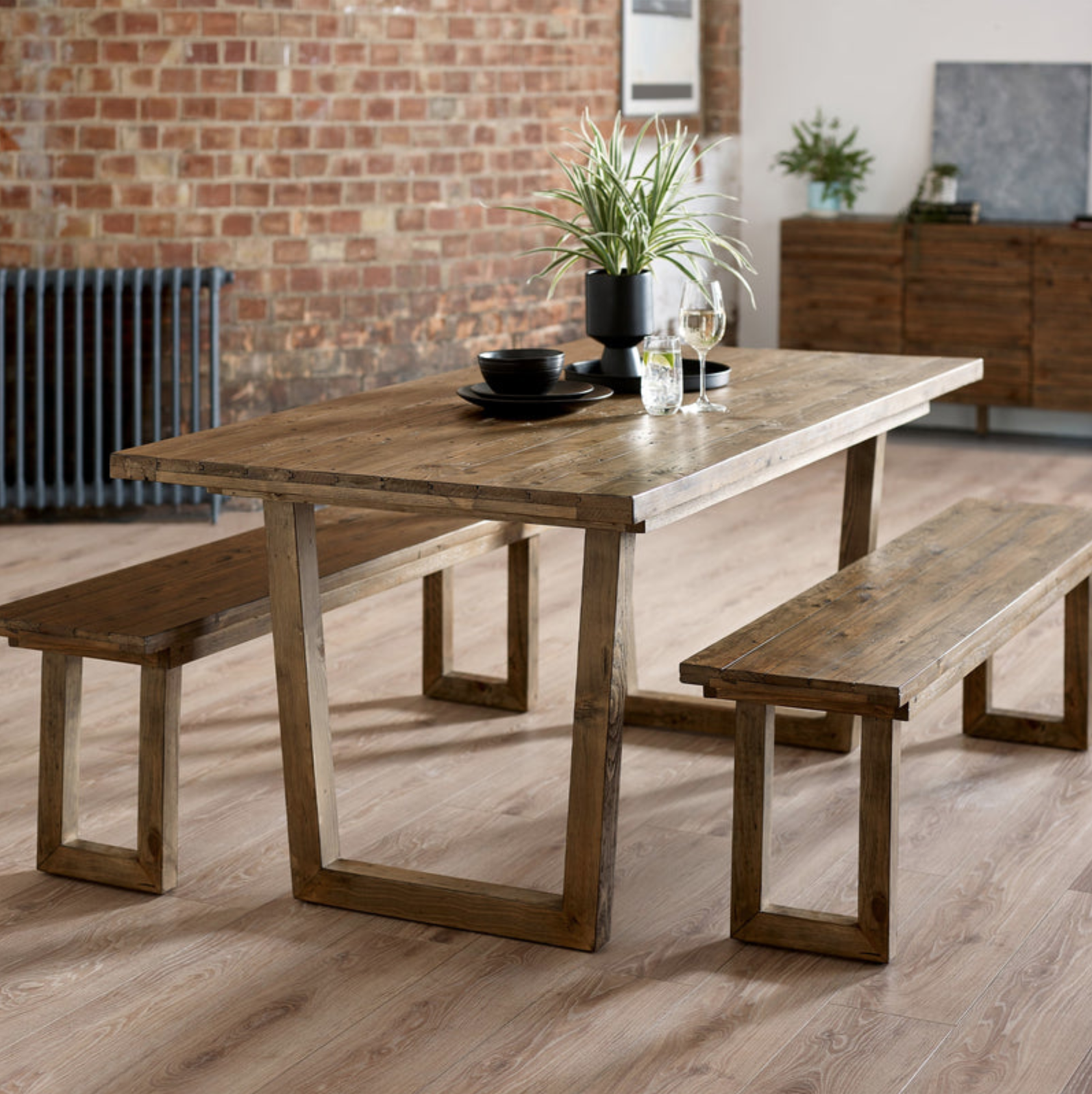 Wooden dining table with two matching benches, decorated with a black vase holding green plants, black bowls and plates, water glasses, and a wine glass in a modern dining area with brick wall and wooden furniture.