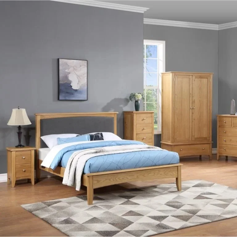 Bedroom with gray walls, wooden bed frame, blue and white bedding, nightstand with lamp, two dressers, wardrobe, and window with sunlight, beige and gray geometric rug on hardwood floor