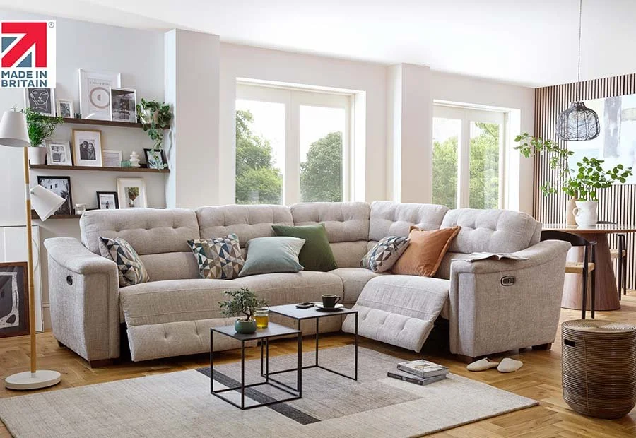 Bright living room with beige sectional sofa, colorful pillows, coffee tables, and large windows showing greenery outside.