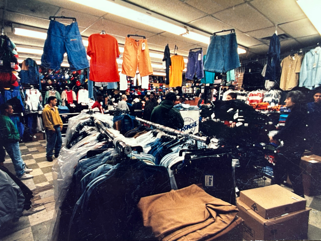 Inside a busy thrift store with clothing on racks and hanging from the ceiling, shoppers browsing through various clothing items and accessories.