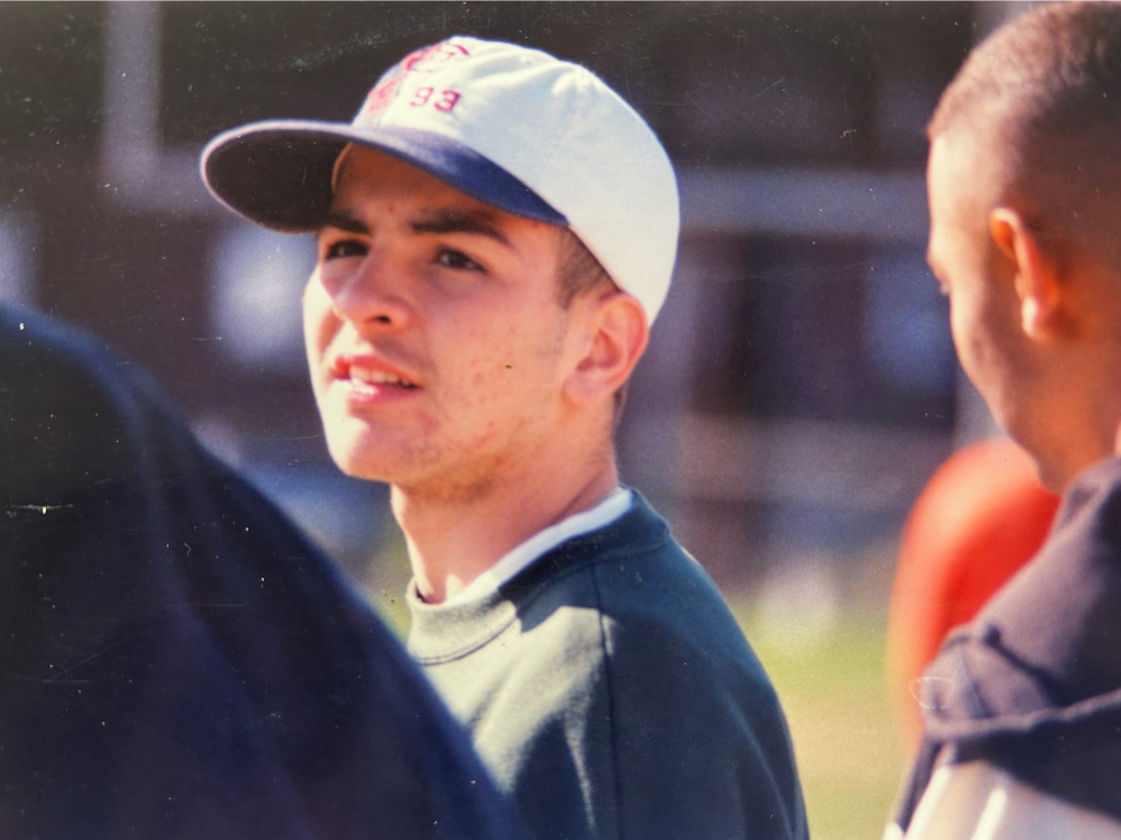 Young man wearing a white cap speaking to a person with short hair dressed in a dark jacket outdoors.