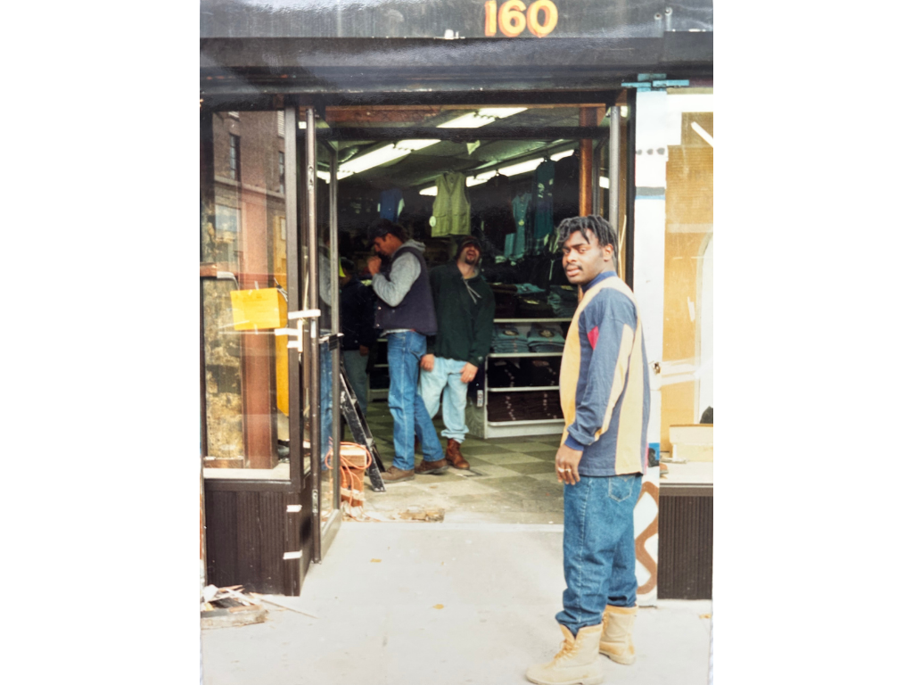 Young man standing outside a store with multiple people inside. The store appears to sell clothing, with shirts and pants visible. The young man is wearing a colorful jacket, jeans, and tan boots, looking at the camera. The store's street number is 1