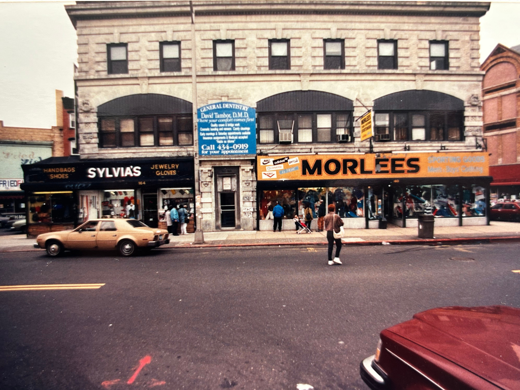 Street view of a two-story brick building with two storefronts: Sylvia's jewelry, gloves, handbags, and shoes, and Morlees sporting goods. Pedestrians are walking on the sidewalk, and a tan vintage car is parked on the street.