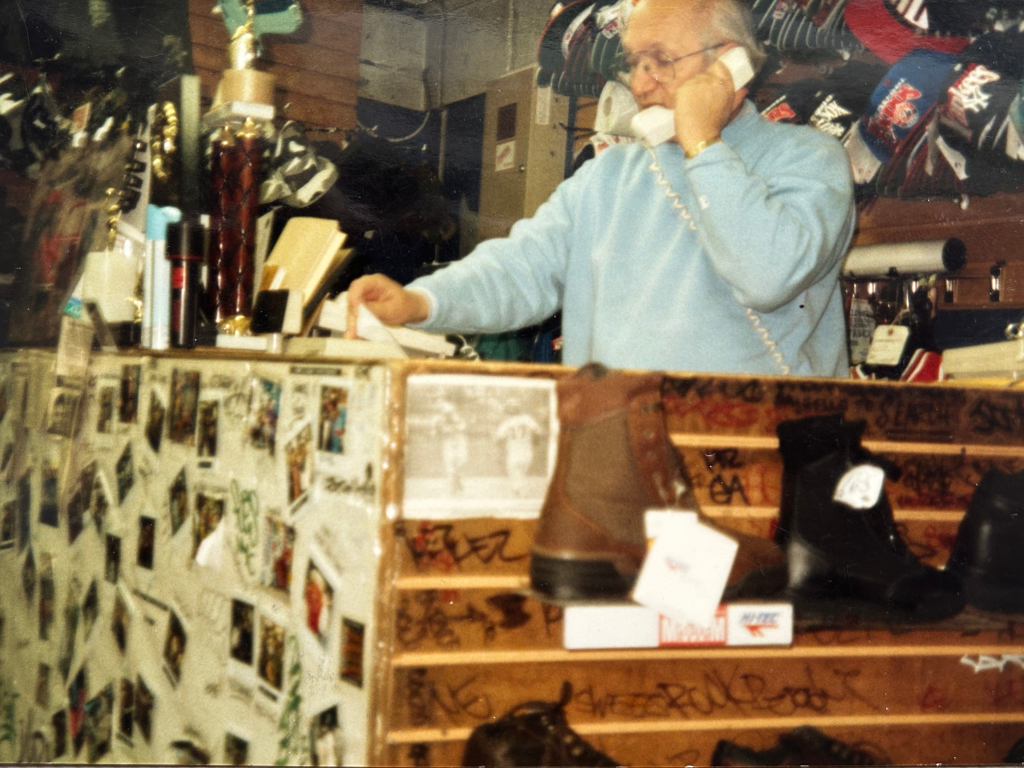 An elderly man with glasses, wearing a light blue sweatshirt, is on the phone at a small shop or store that sells shoes and trophies, with various items and photographs displayed around him.