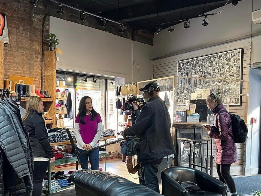 A female interviewee speaking to a camera crew inside a retail store, with clothing and shoes displayed on shelves and walls.