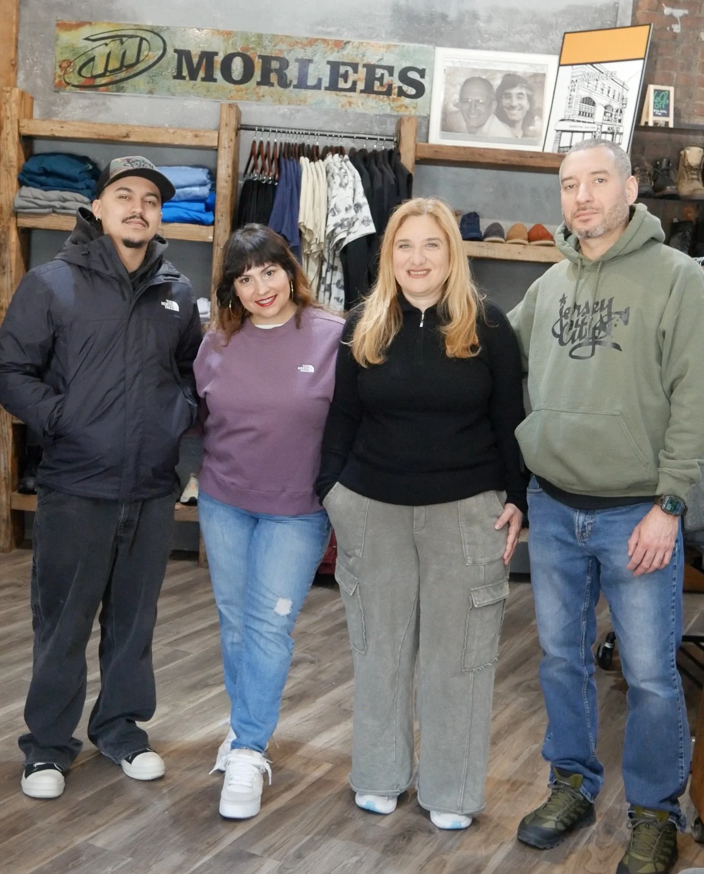 Four people standing inside a clothing store, smiling, with shelves of clothes, shoes, and framed photographs in the background.