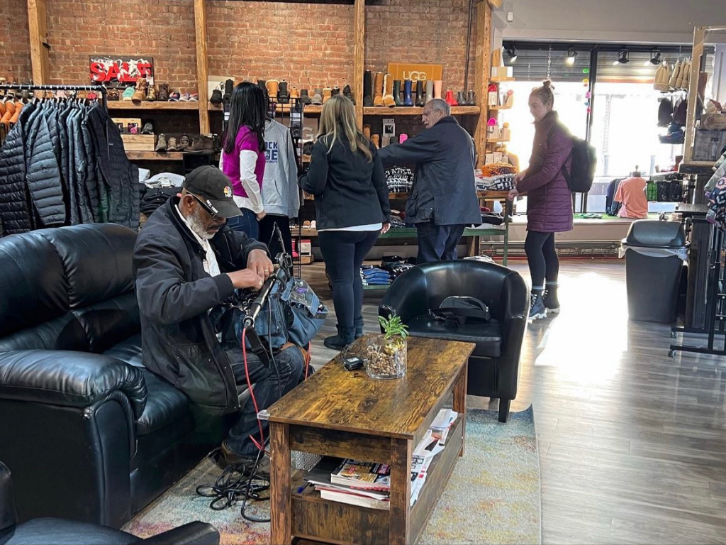 People shopping in a retail store with jackets, boots, and clothing on display, and a man sitting on a black leather couch.