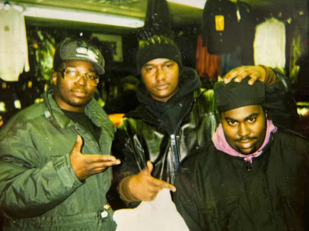 Three young men standing indoors, two with hats, wearing dark jackets, with serious expressions, and making hand gestures.