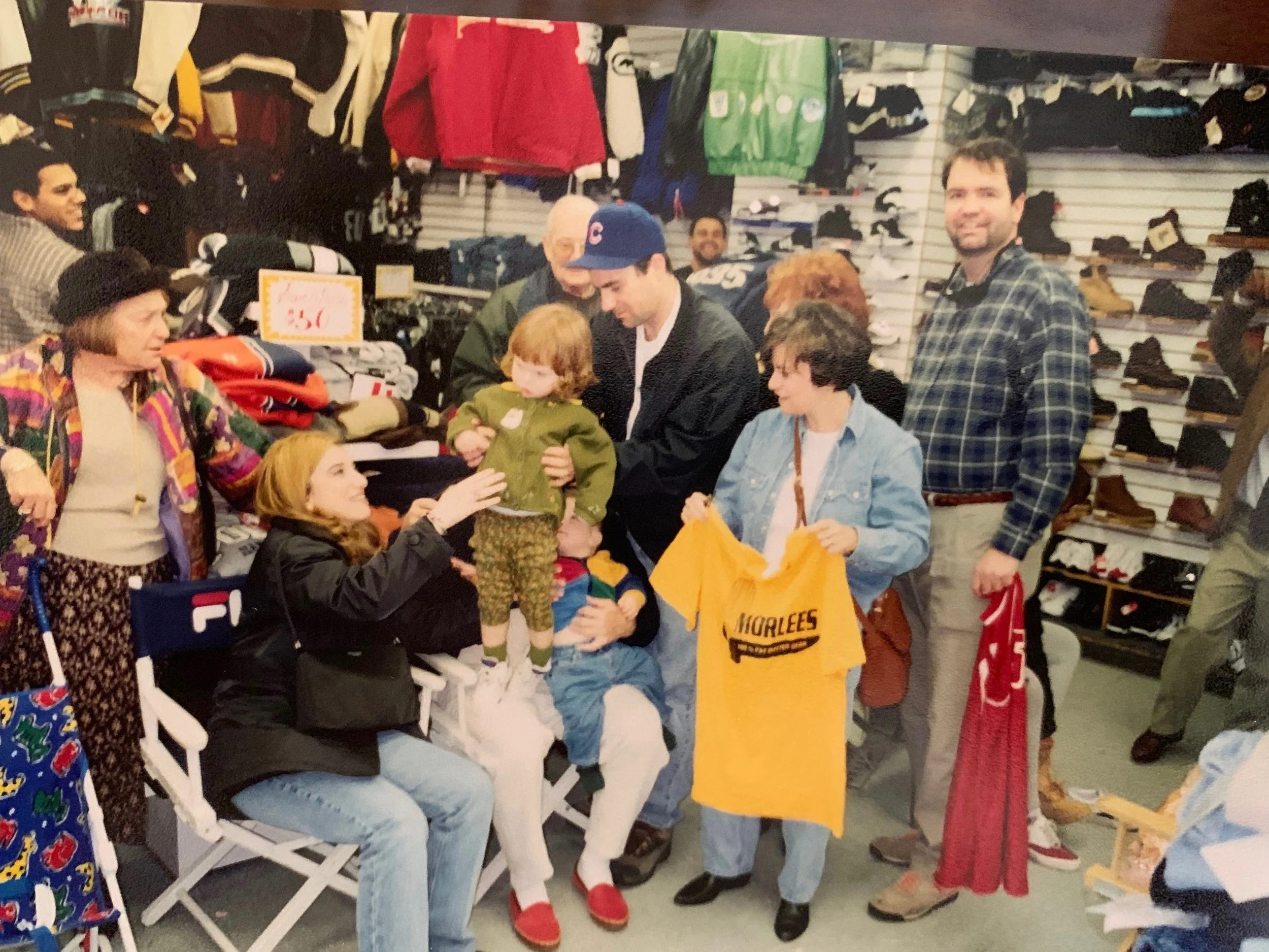 A group of people shopping at a sporting goods store counter, with a young girl on someone's lap and a woman holding a yellow T-shirt, amidst shelves of shoes and sports gear.