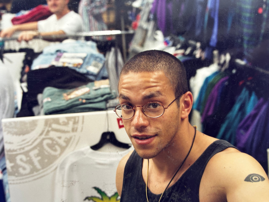 A young man with glasses, a Tattoo on his left arm, and wearing a black tank top, taking a selfie inside a clothing store with racks of colorful clothes and folded garments in the background.